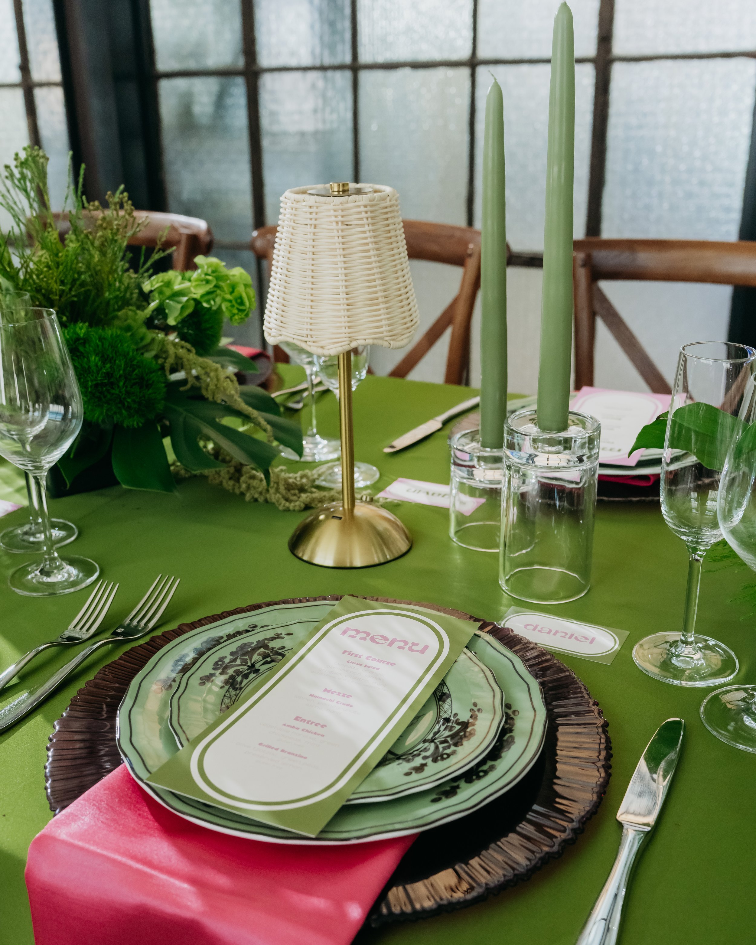A table setting with a green tablecloth, various glassware including wine and water glasses, a decorative table lamp with a woven lampshade, two tall green candles in glass holders, fresh green leaves and flowers, and a menu card placed on a decorati