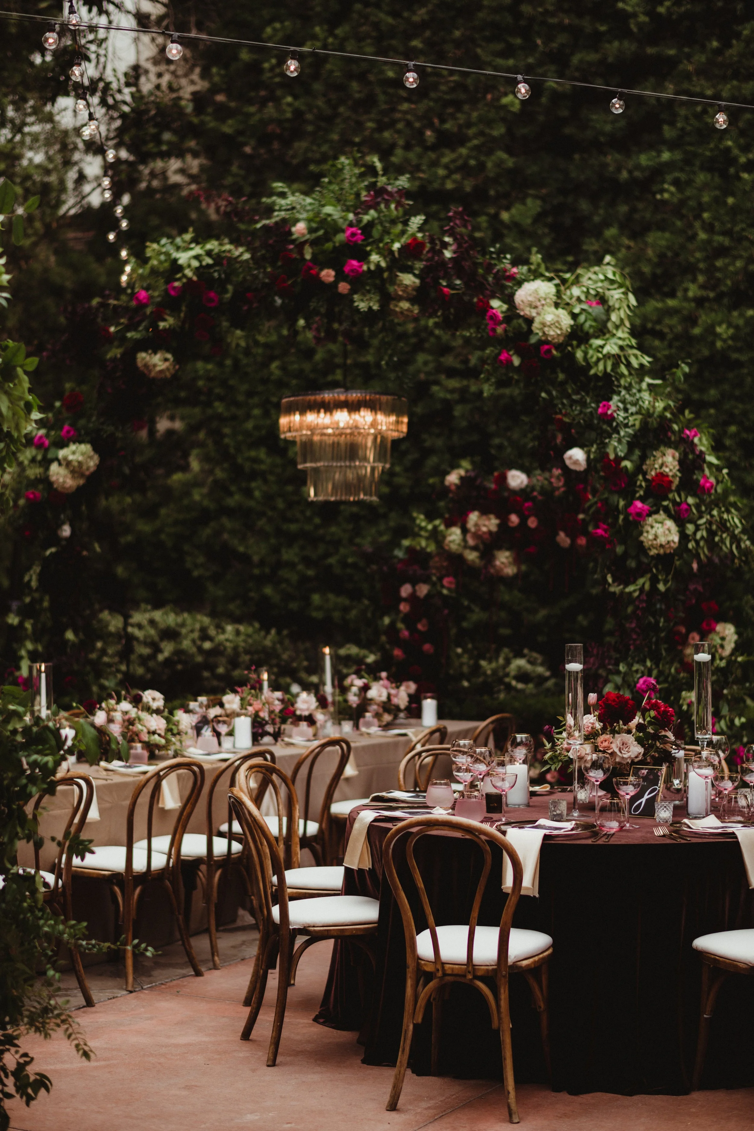 Elegant outdoor wedding reception setup with a long banquet table decorated with pink, white, and red floral arrangements, candleholders, and glassware under string lights and a floral arch surrounded by greenery.