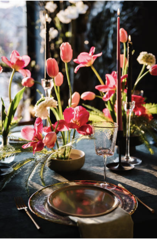 Elegant table setting with pink and white flowers, tall pink taper candles, a wine glass, and a decorative plate with a napkin, on a dark table.