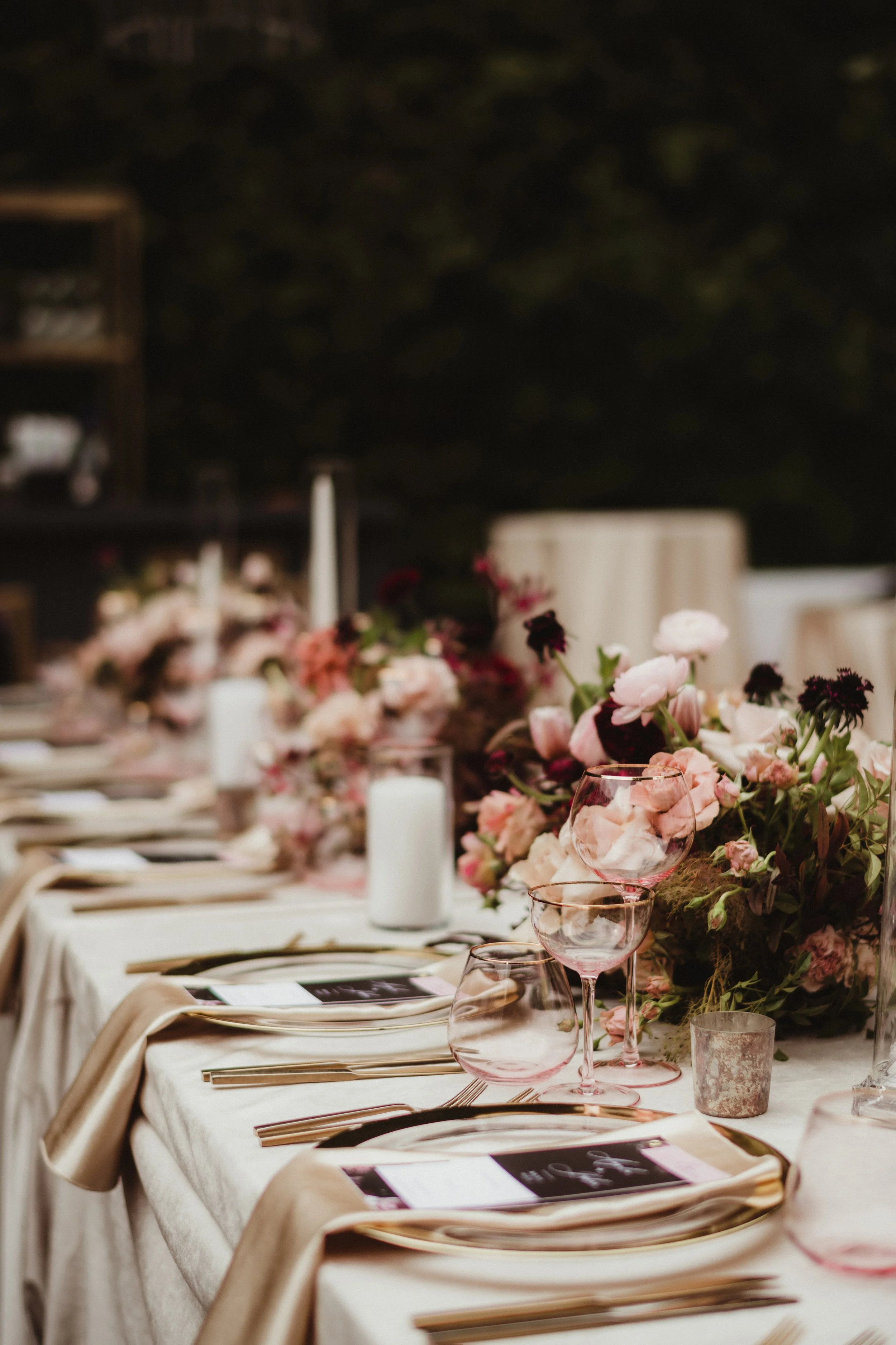 A table set for a formal event with pink and dark red flowers and pink wine glasses, silverware, napkins, and menus on a cream-colored tablecloth.