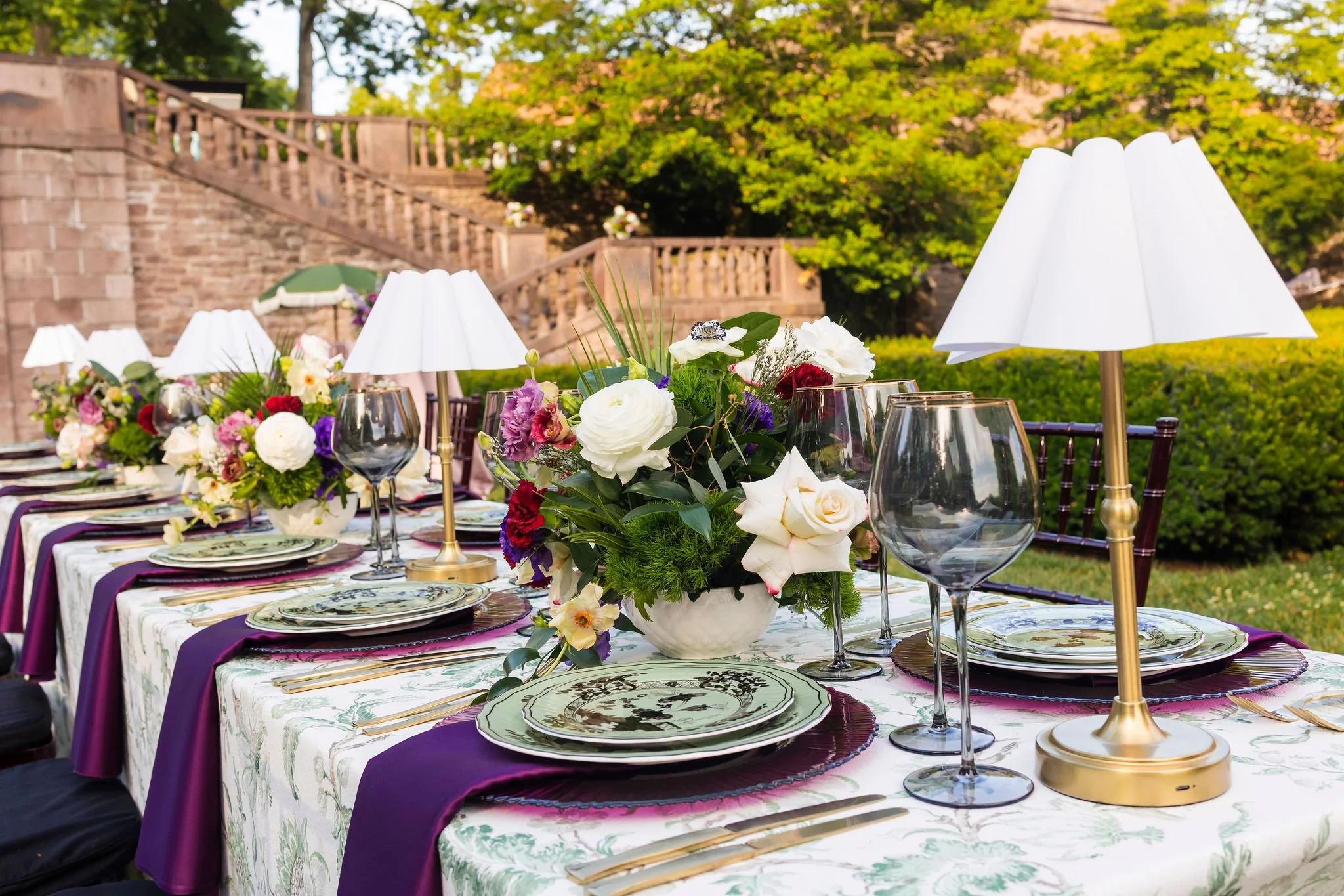 Outdoor dining table decorated with floral arrangements, tableware, glasses, and white lampshades, with greenery and a brick staircase in the background.