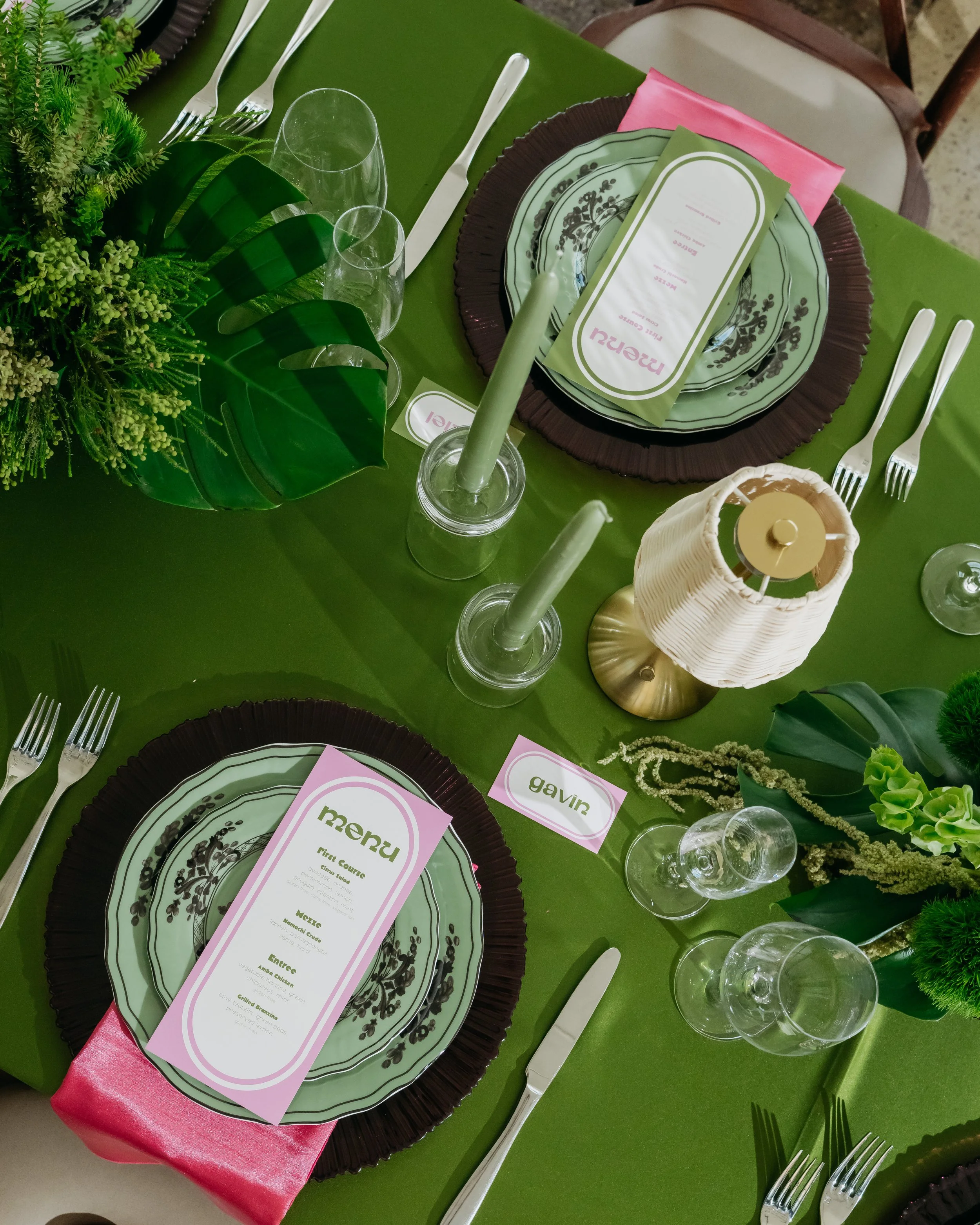 A table setting with green tablecloth, decorative plates, glassware, and utensils. There are green foliage and floral arrangements, pink napkins, and place cards with names on the table.