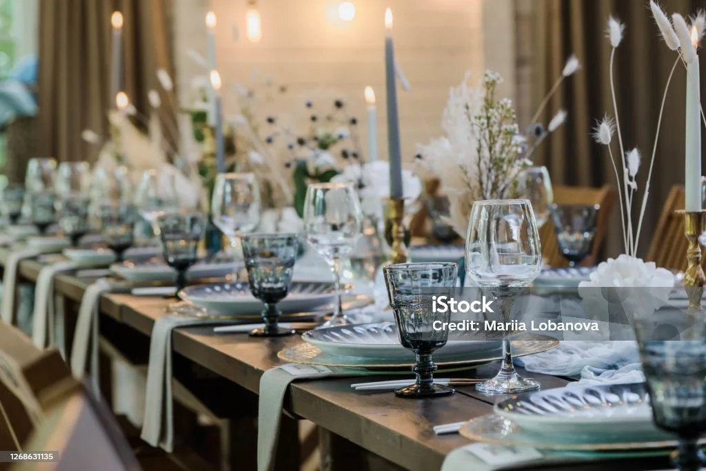 A long dining table set for a formal event with glasses, plates, napkins, and floral centerpieces in a decorated room.