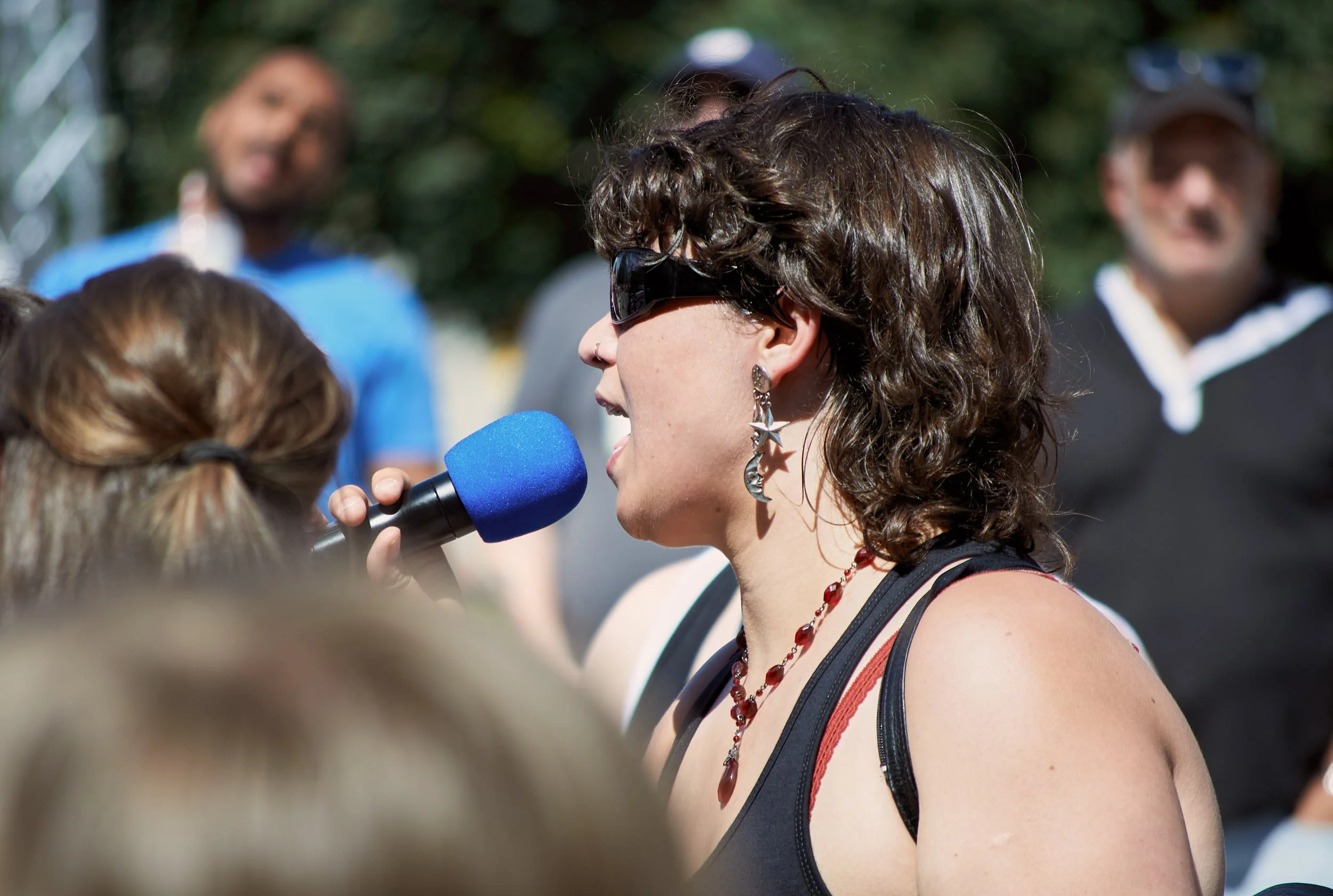A woman wearing dark sunglasses and star-shaped earrings is speaking into a microphone during an outdoor event with a group of people in the background.