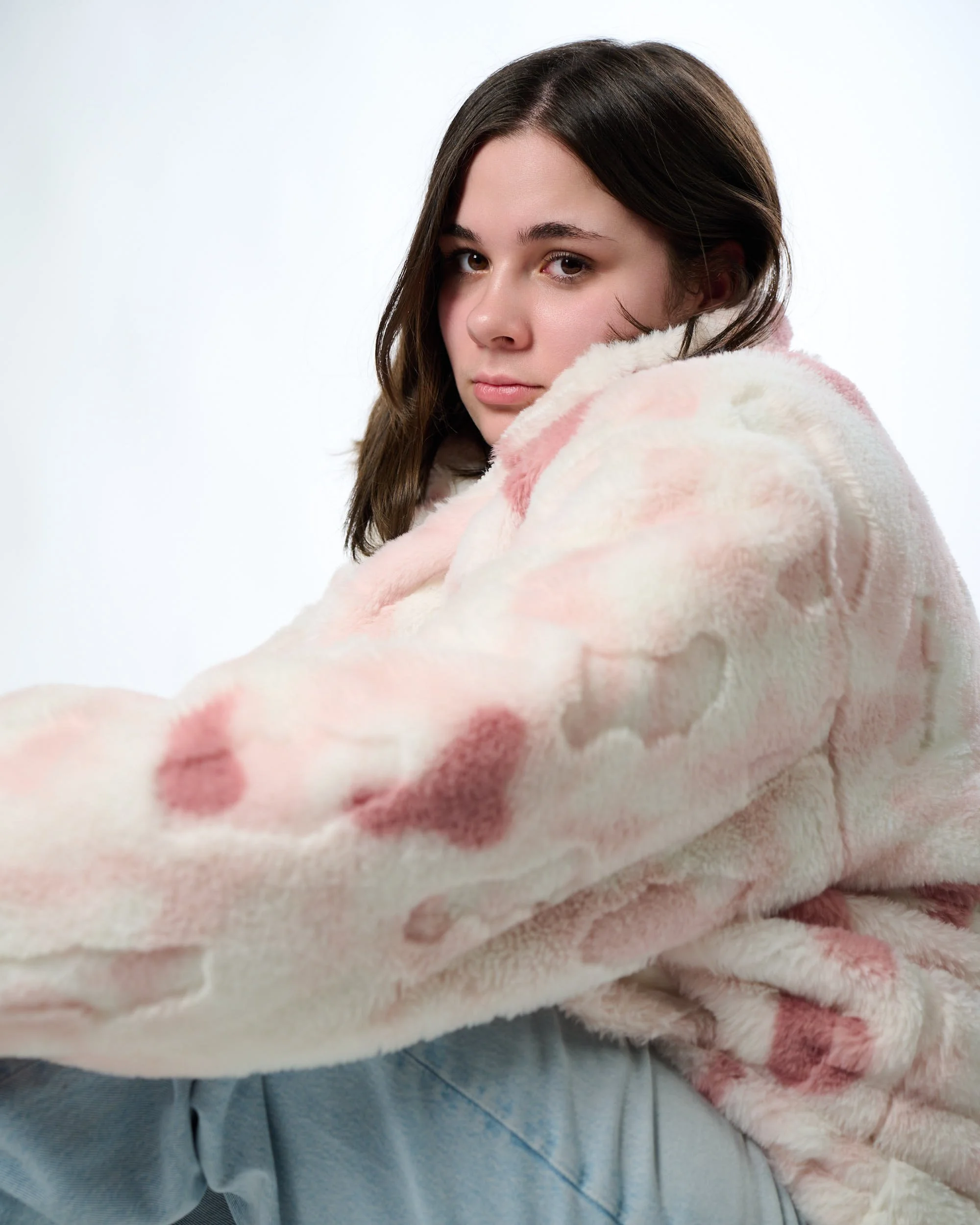 A young woman with brown hair wearing a pink and white plush jacket with red heart patterns, looking at the camera against a plain white background.
