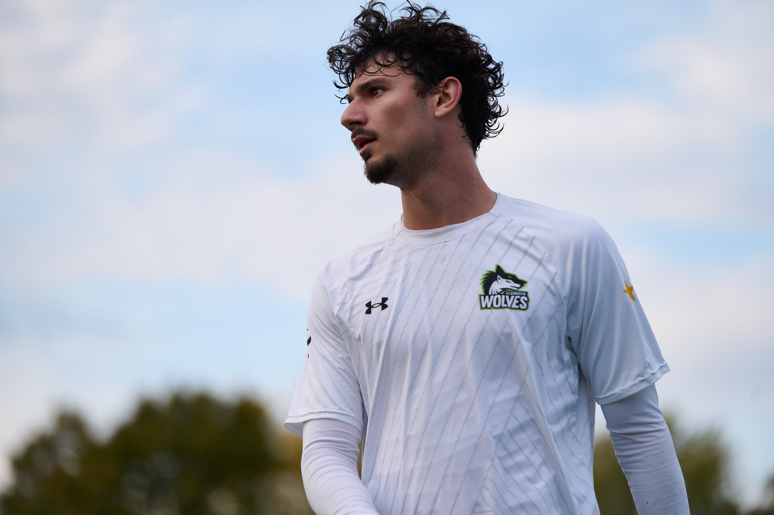 A male soccer player with curly dark hair and light skin, wearing a white Alouquin Wolves jersey, standing outdoors against a cloudy sky with blurred trees in the background.