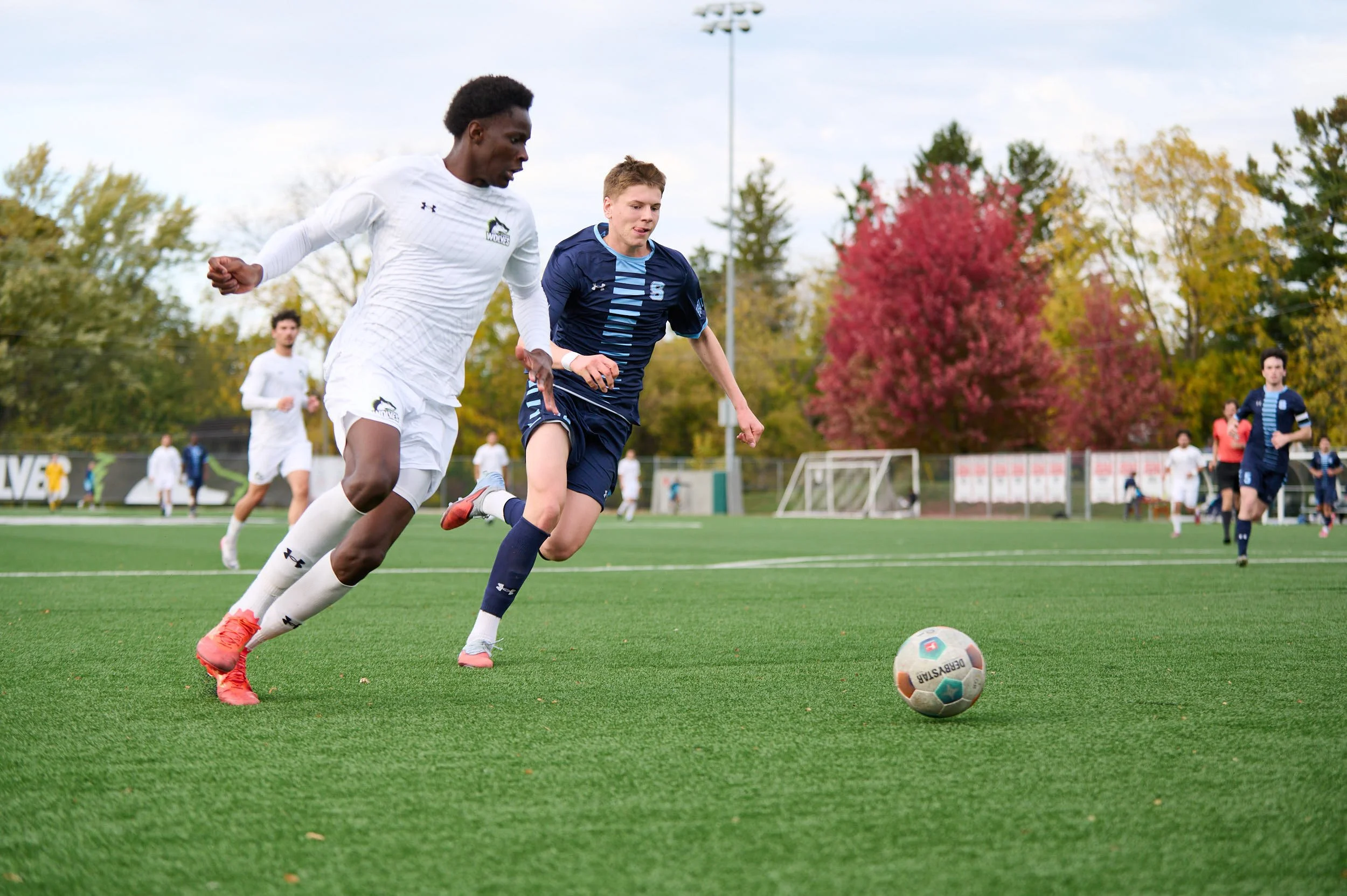 Two young male soccer players competing for the ball on a green field during a match, with other players and trees with fall foliage in the background.