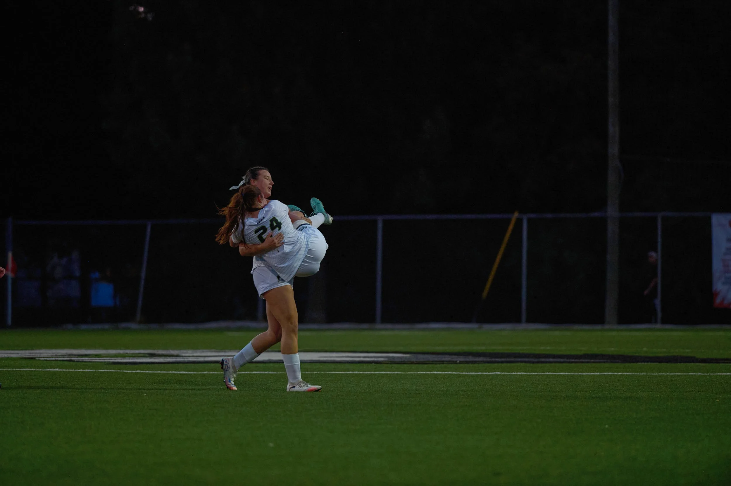 Two female soccer players in white jerseys, one lifting the other in celebrating on the field at night.