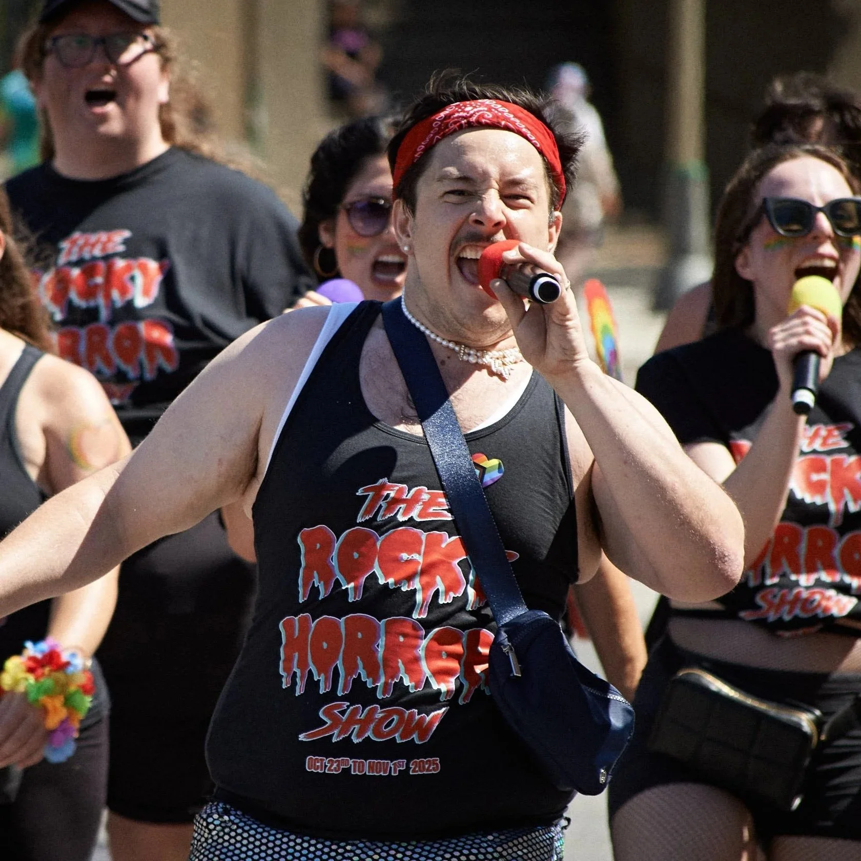 People participating in a pride parade, wearing black tank tops with 'The Rock Horror Show' logo, some holding rainbow-colored items, and one person singing into a microphone with a red headband and a pearl necklace.