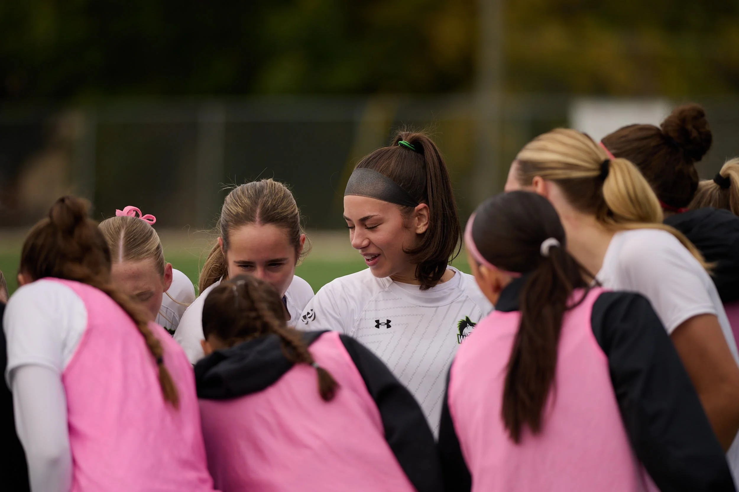 A group of young female soccer players huddled together in a circle on a field, listening to their coach or team leader.