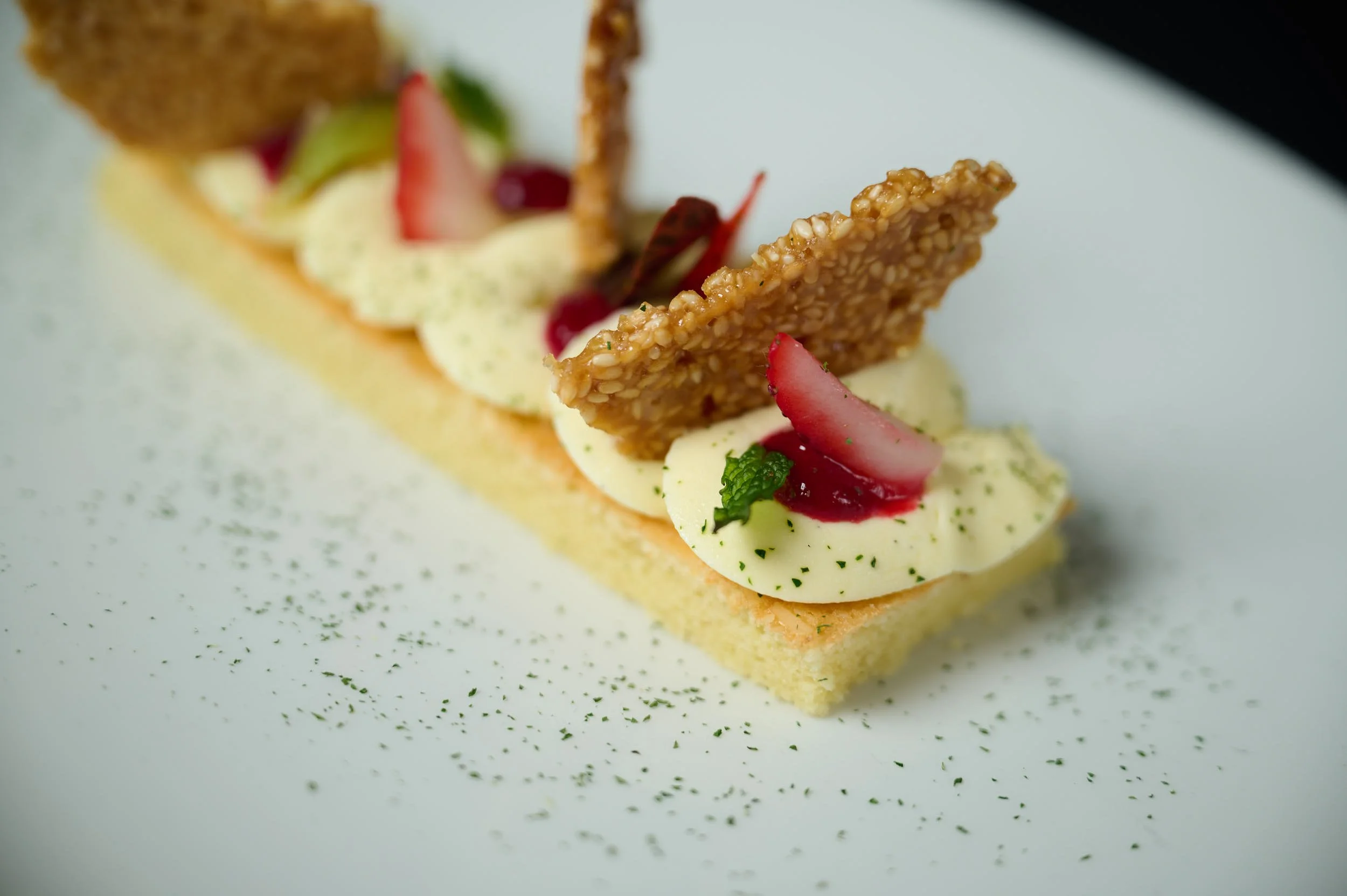 A rectangular dessert topped with dollops of cream, sliced strawberries, a mint leaf, and a sesame crispy biscuit, garnished with green herbs on a white plate.