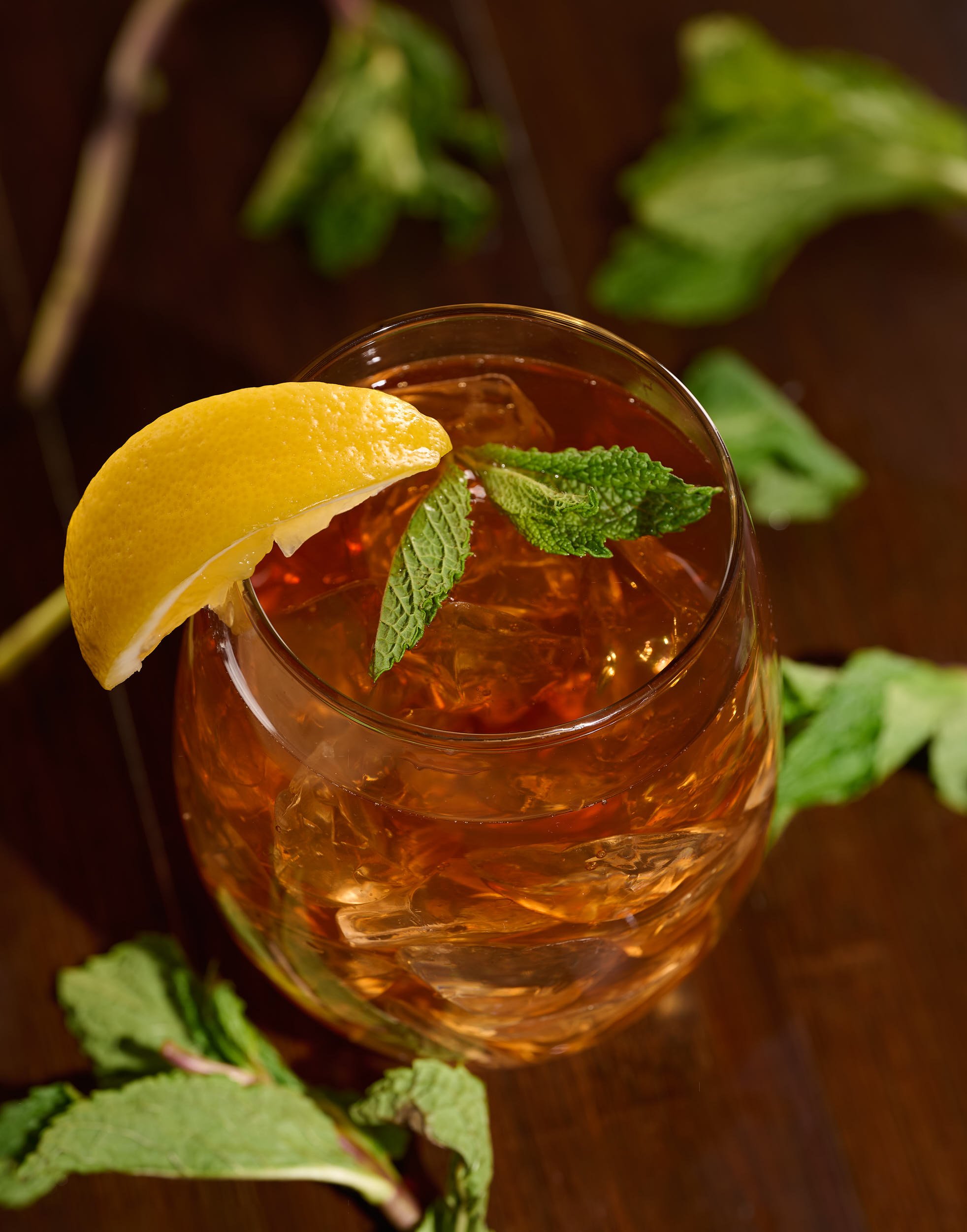 A glass of iced tea garnished with a lemon wedge and fresh mint leaves, placed on a wooden surface with scattered mint leaves around.