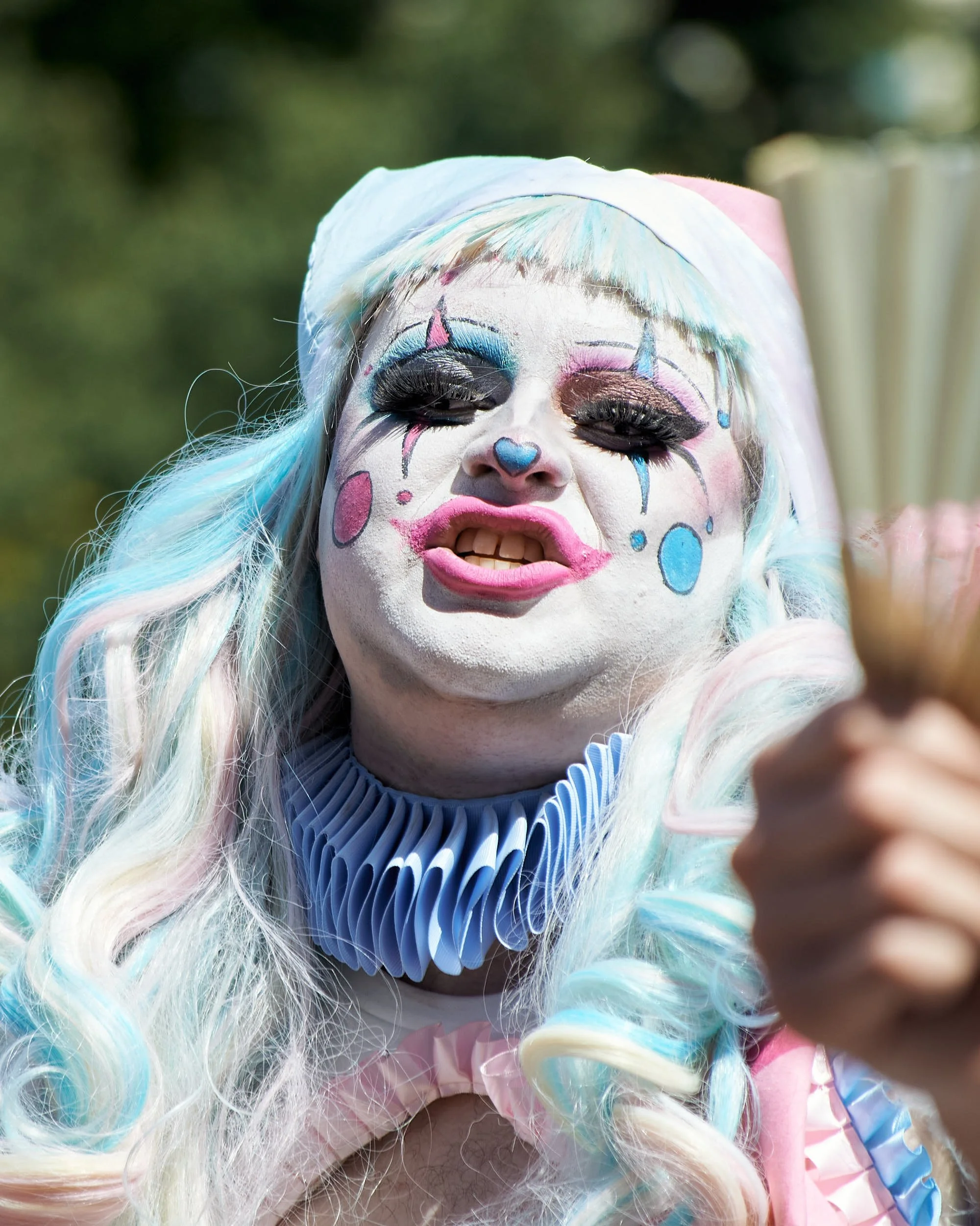 Person dressed as a clown with white face paint, colorful makeup, blue and pink hair, wearing a ruffled collar, and holding a fan.