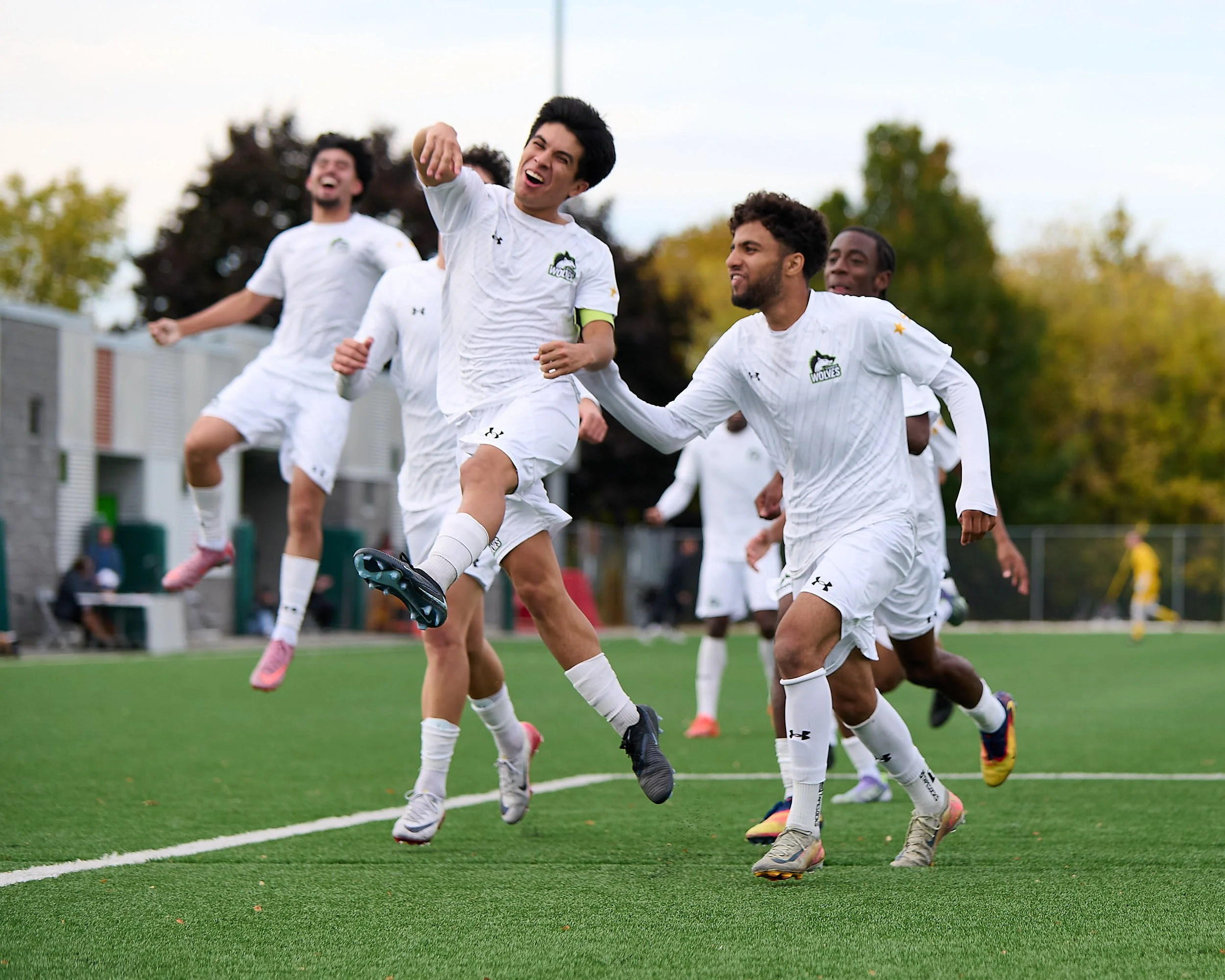 Soccer players in white jerseys celebrating on the field, jumping and running, with trees and a building in the background.