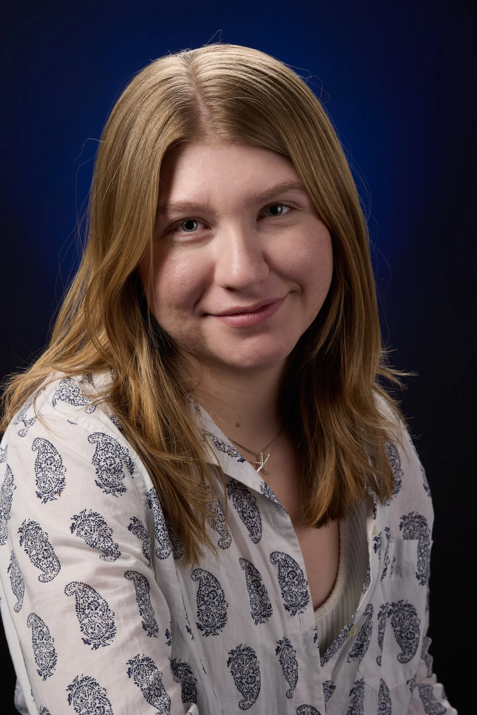 Portrait of a young woman with shoulder-length, light brown hair, wearing a white shirt with a blue paisley pattern, and a silver necklace with a letter 'K' pendant, against a dark gradient background.