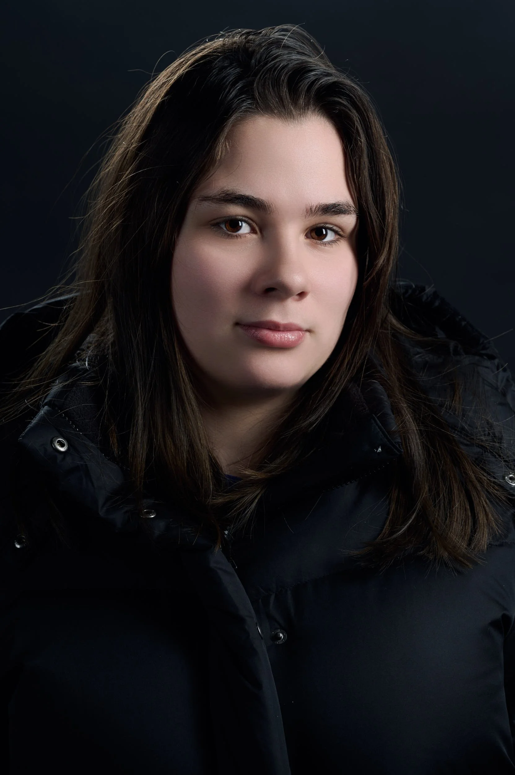 Portrait of a young woman with brown hair wearing a black jacket against a dark background.