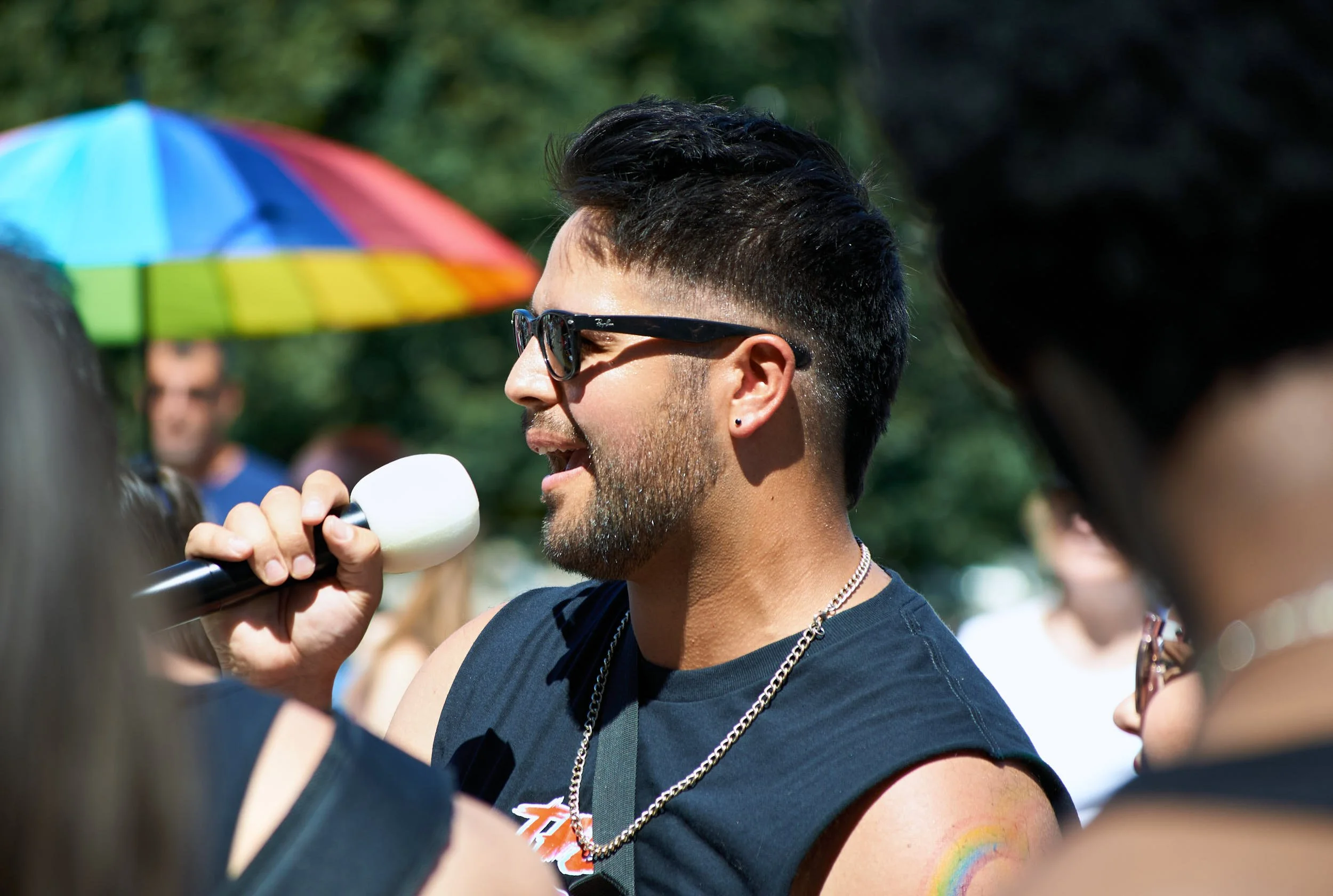 A man with sunglasses and a beard speaking into a microphone at an outdoor event, with a rainbow tattoo on his arm, and colorful umbrella and several people in the background.