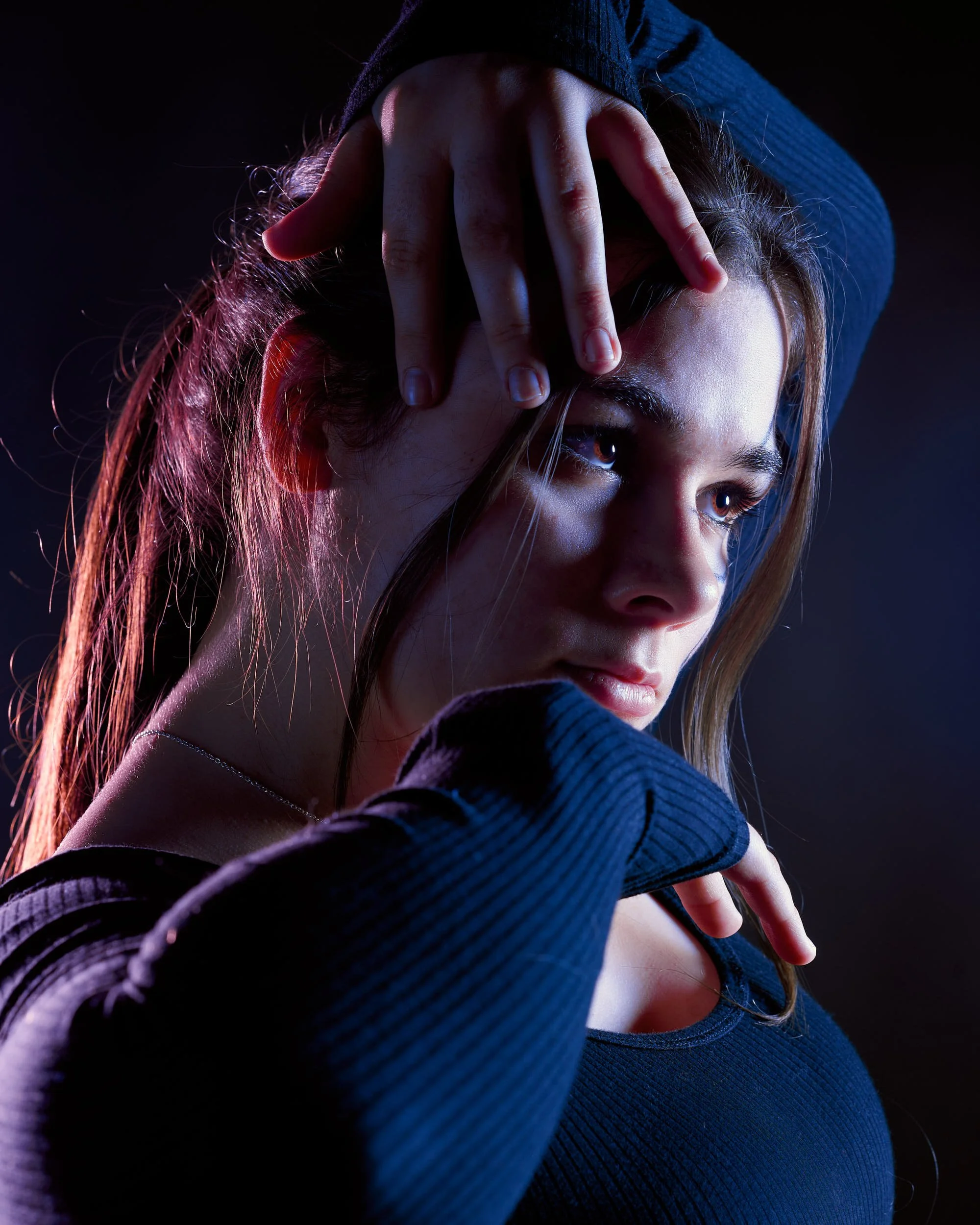 Close-up of a young woman with long hair, wearing a dark long-sleeve shirt, looking thoughtfully into the distance with her hand gently touching her face and head against a dark background.