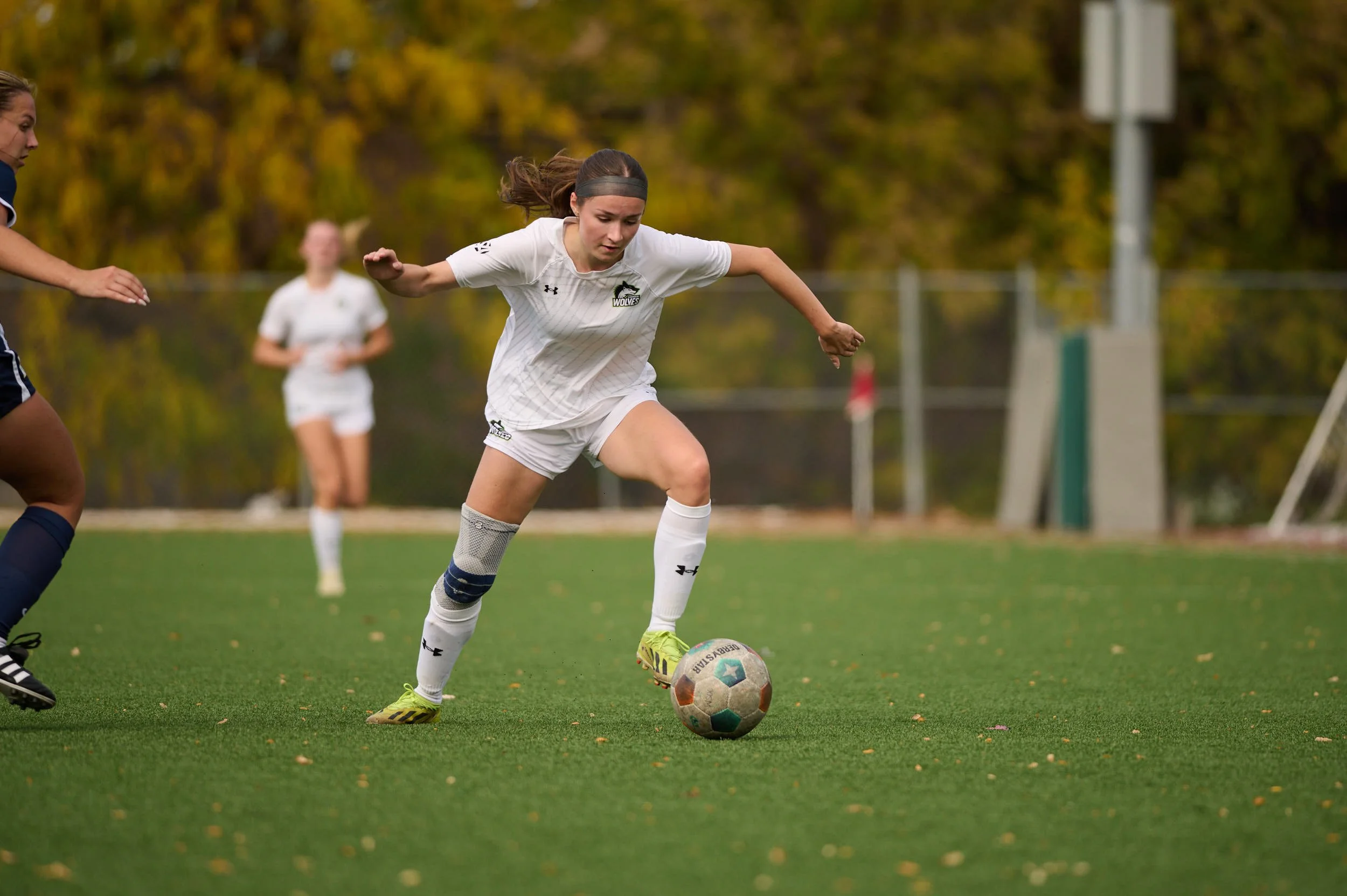 Female soccer player in white uniform dribbling a soccer ball during a game on a green field with autumn trees in the background.