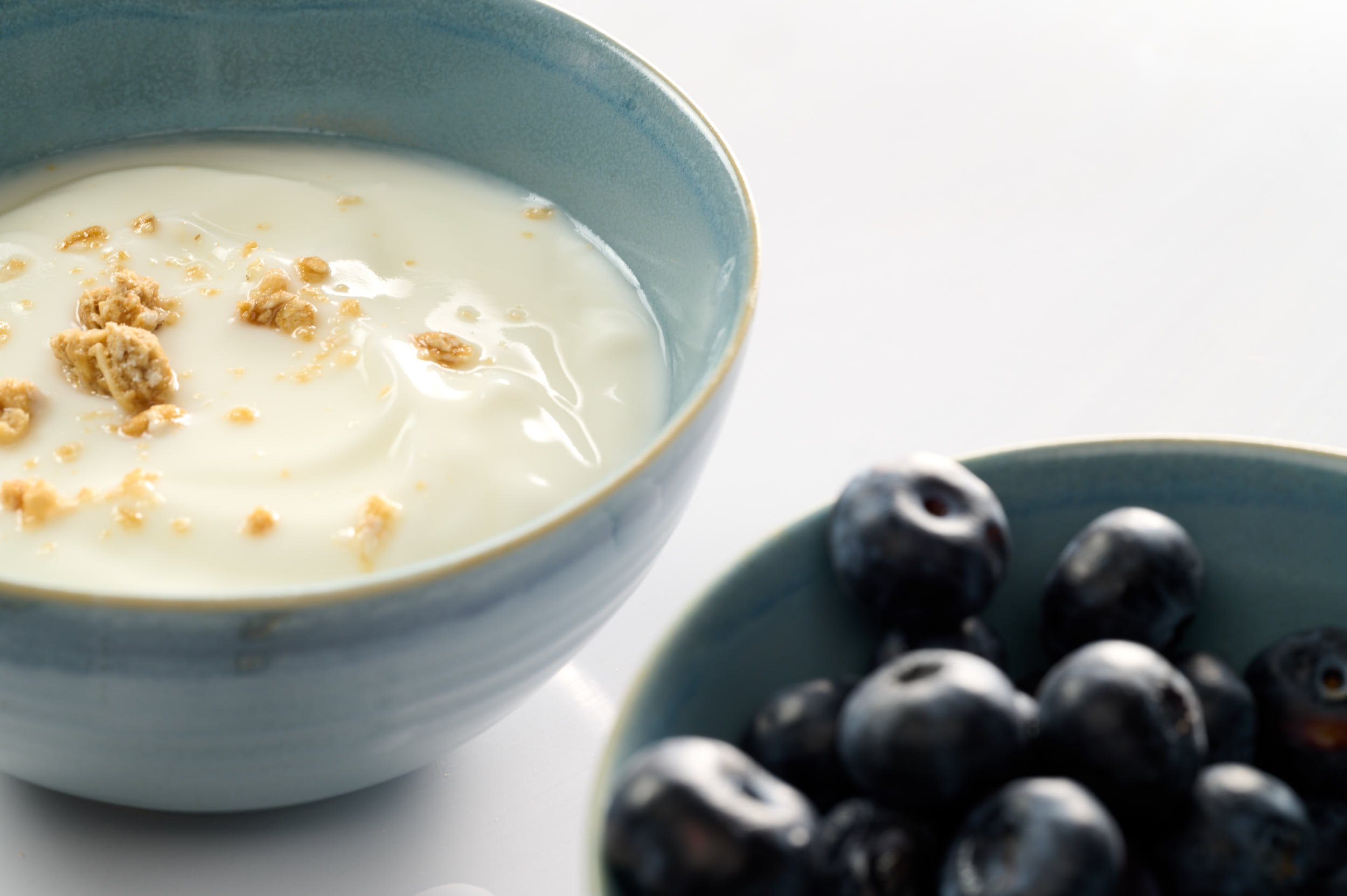 A blue bowl filled with yogurt topped with granola, next to a smaller bowl of fresh blueberries on a white surface.
