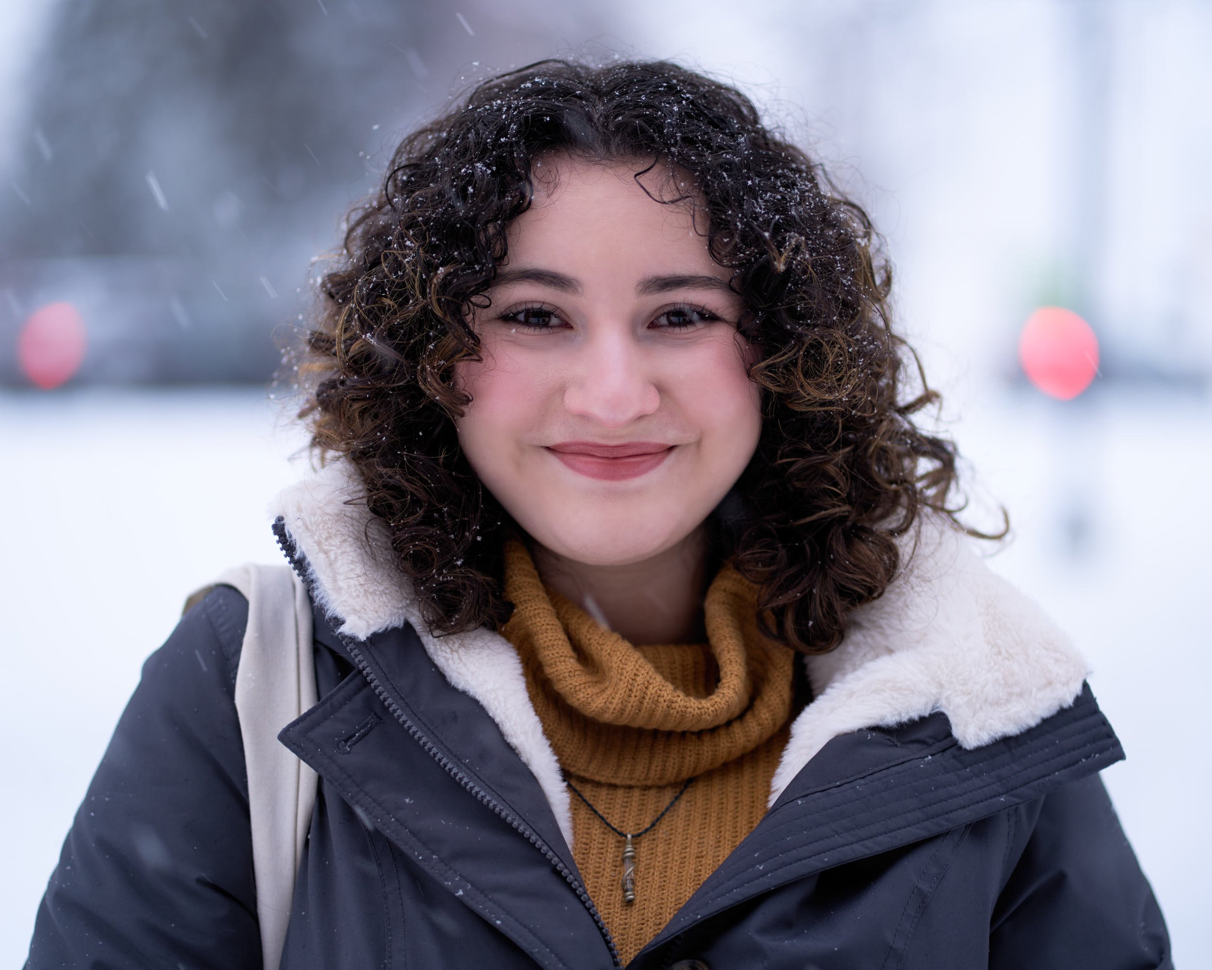 A woman with curly hair smiling outdoors in winter, wearing a dark jacket with a white fleece lining and a mustard-colored turtleneck.