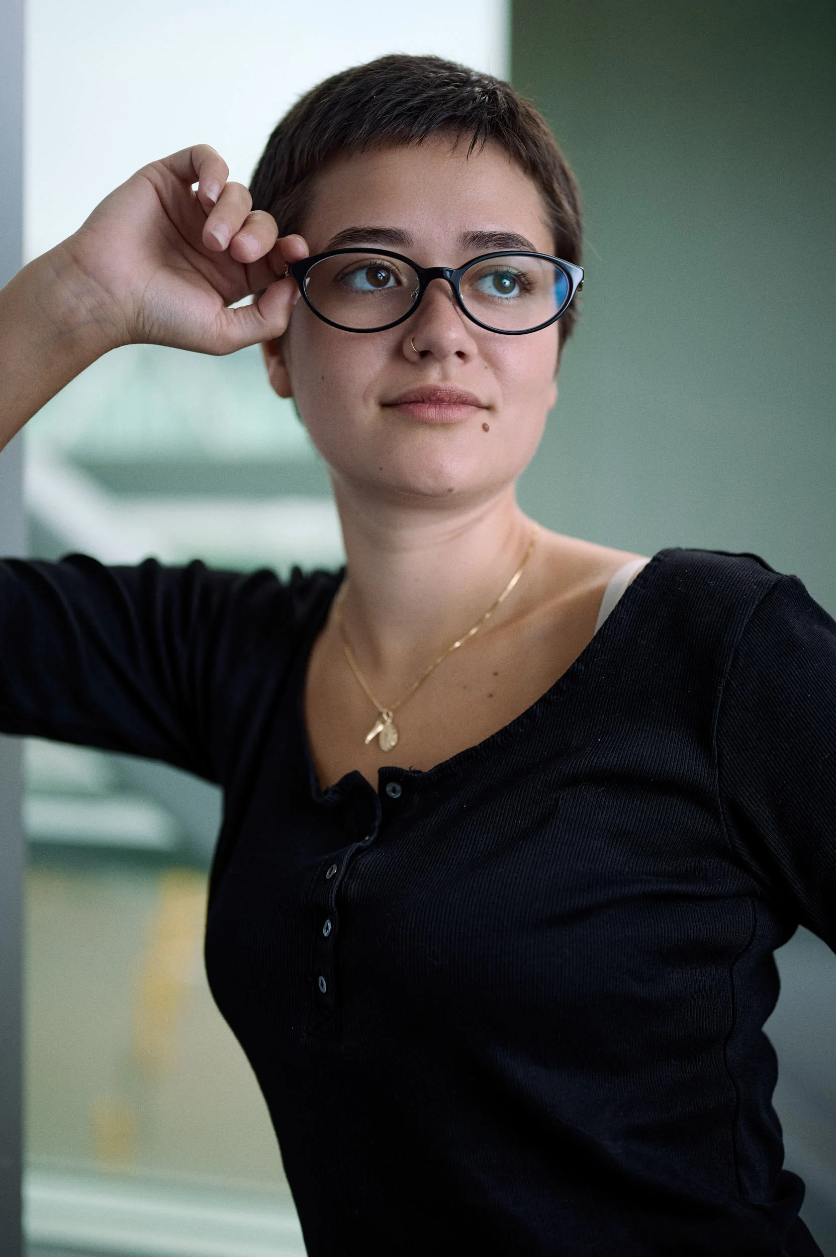 A young woman wearing glasses, a black button-up shirt, a white tank top underneath, a gold necklace, and a small nose ring, adjusting her glasses while standing indoors with a neutral background.