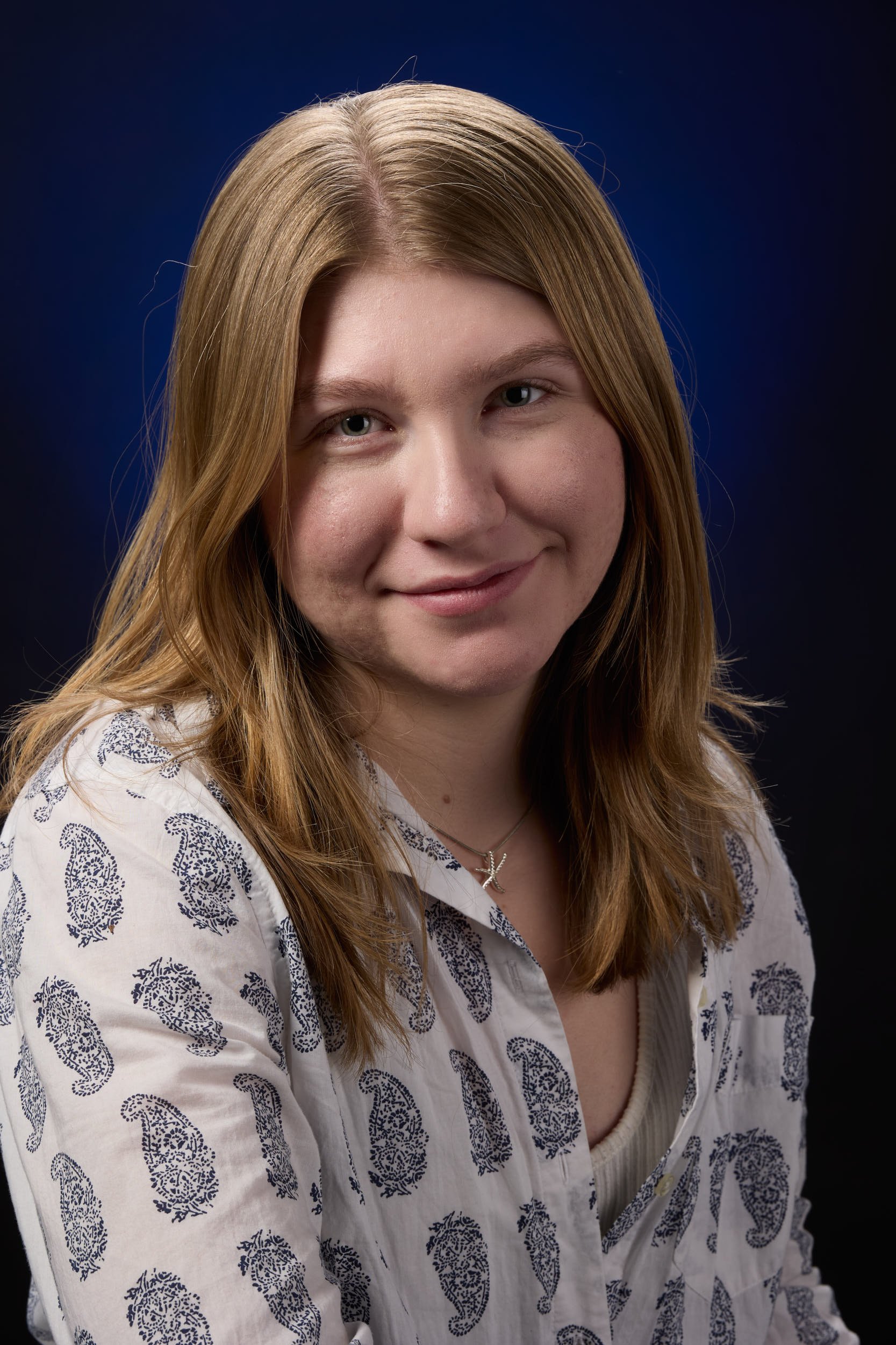Portrait of a young woman with shoulder-length, light brown hair, wearing a white shirt with a paisley pattern, and a silver necklace with a pendant, against a dark blue background.