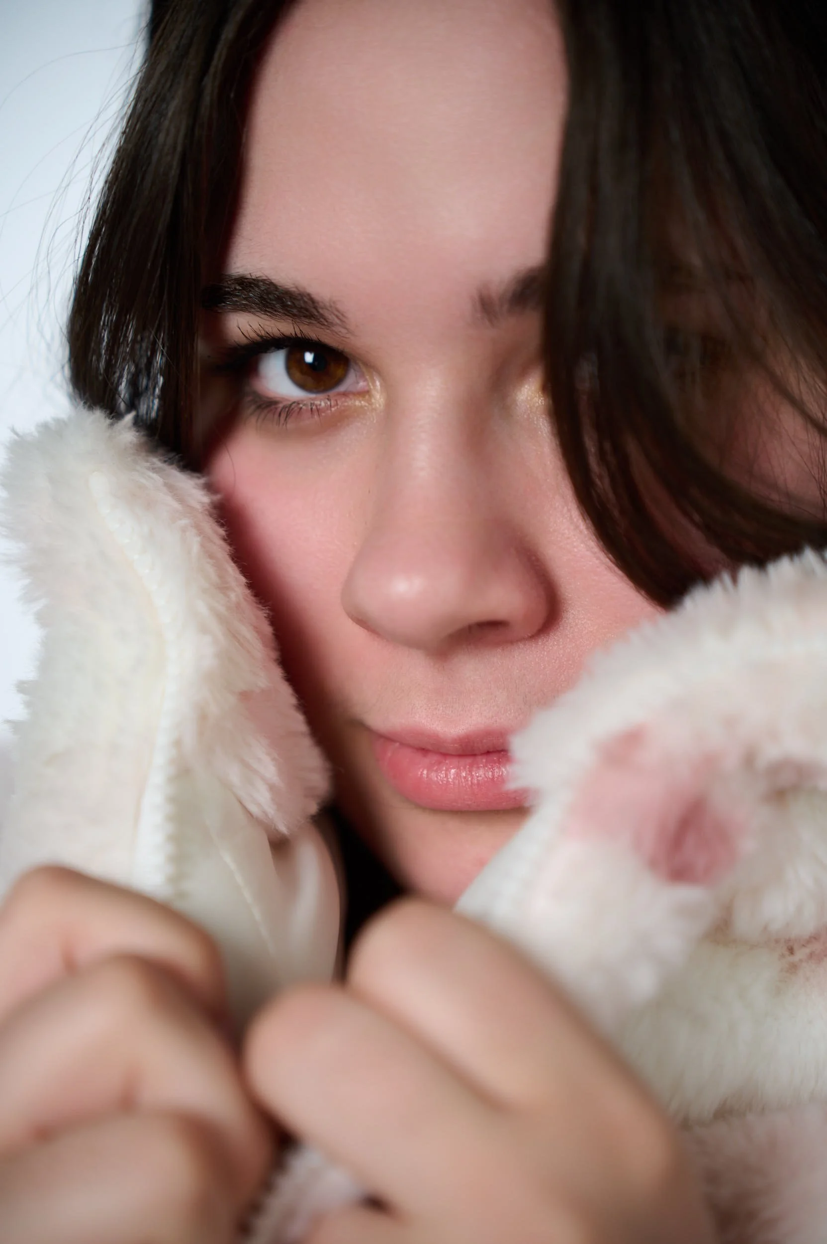 A close-up of a woman with brown hair and brown eyes, holding fluffy white fabric close to her face, looking at the camera.