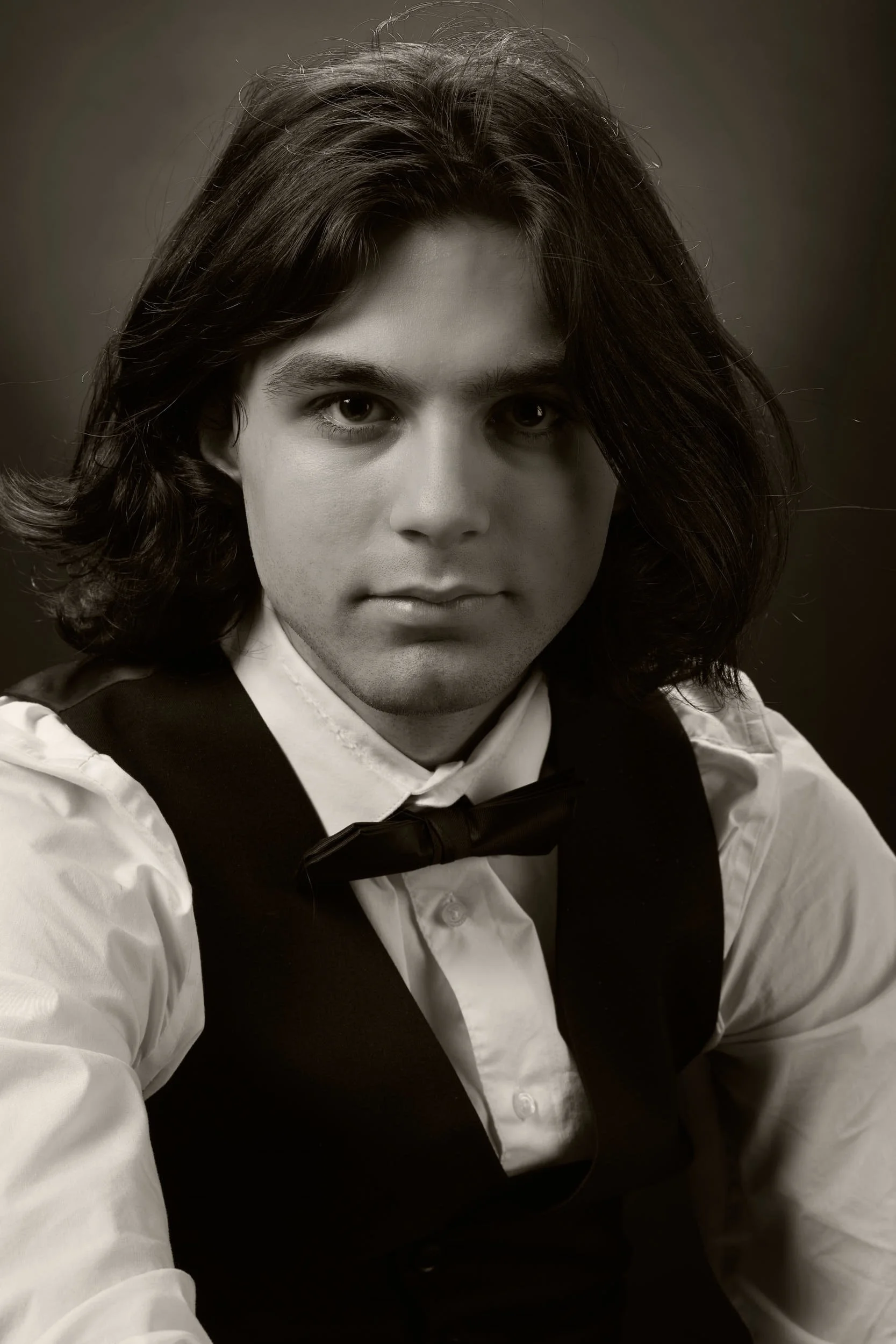 Black and white portrait of a young man with shoulder-length hair, wearing a white shirt, black vest, and a bow tie.