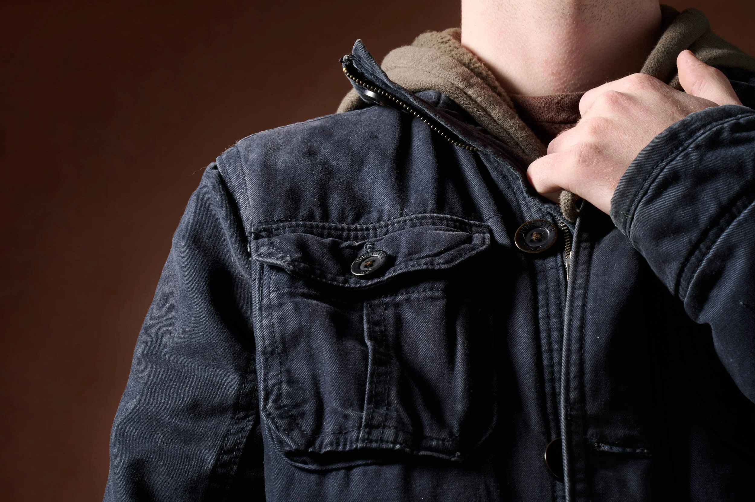 Person adjusting the collar of a dark denim jacket with a brown hoodie underneath and a brown background.