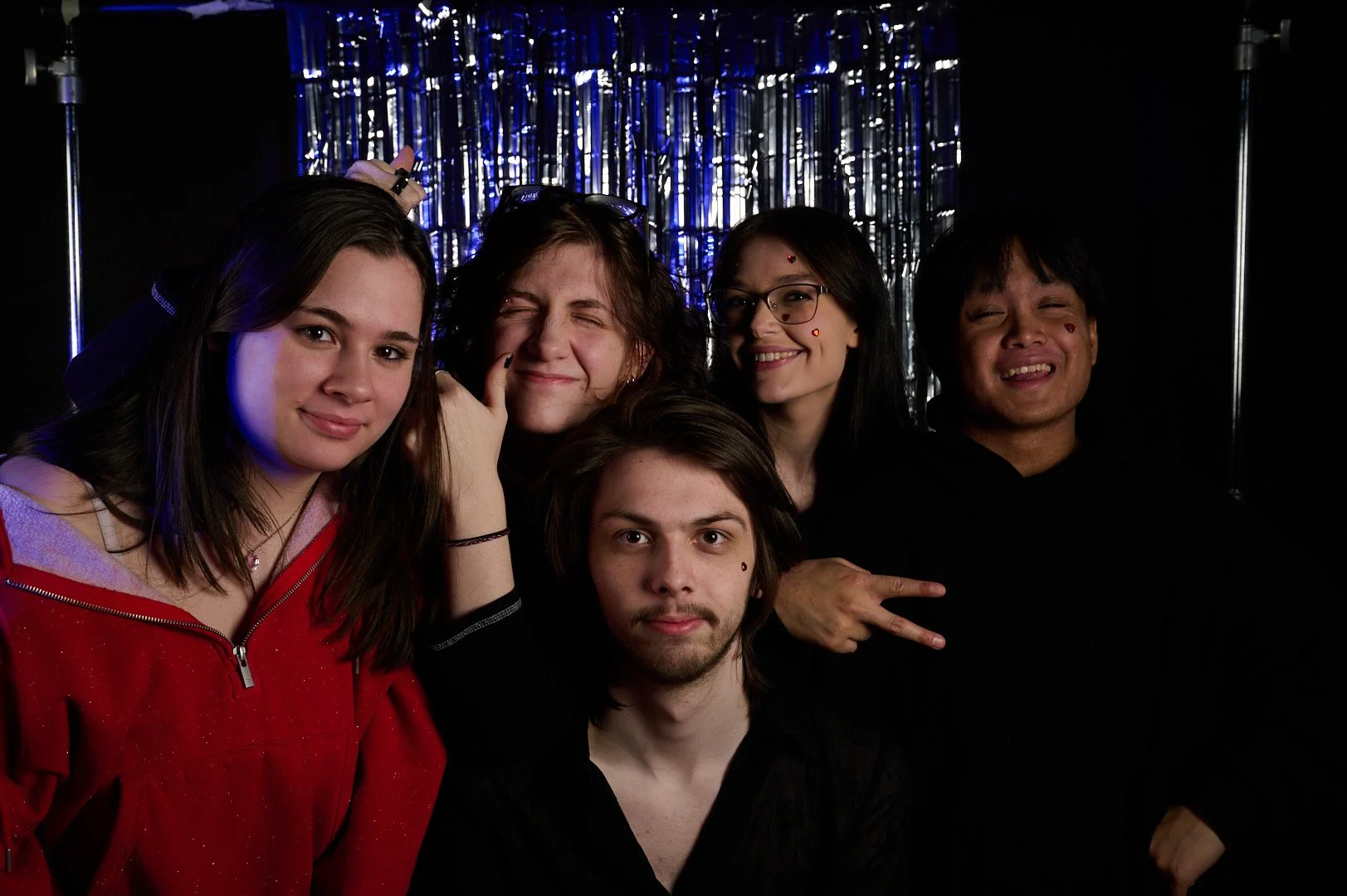A group of five young adults posing together in front of a shiny blue foil backdrop at a party or celebration, with some making playful gestures and expressions.