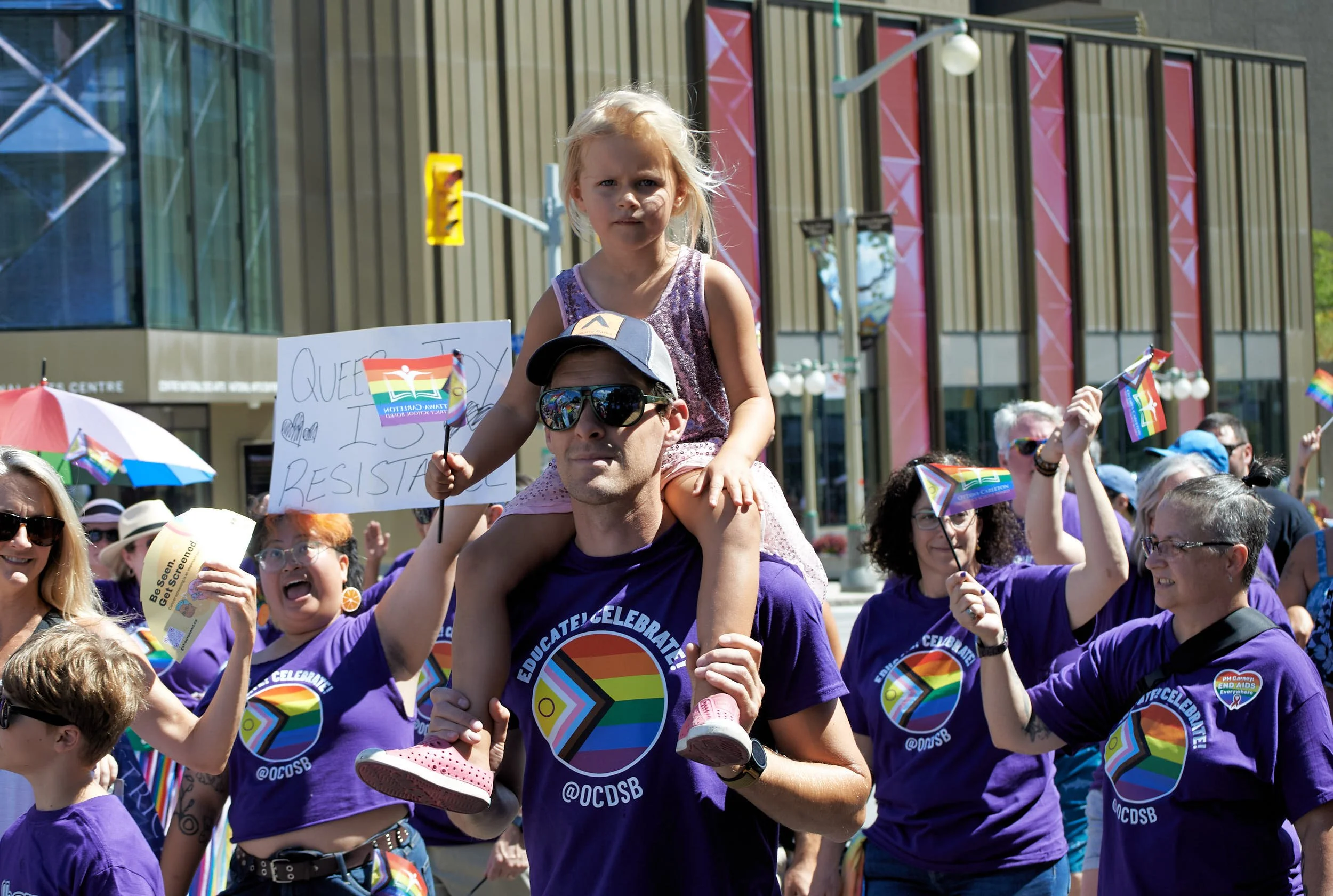 People participating in a Pride parade, wearing purple shirts with rainbow logos, holding rainbow flags, and celebrating on a city street.