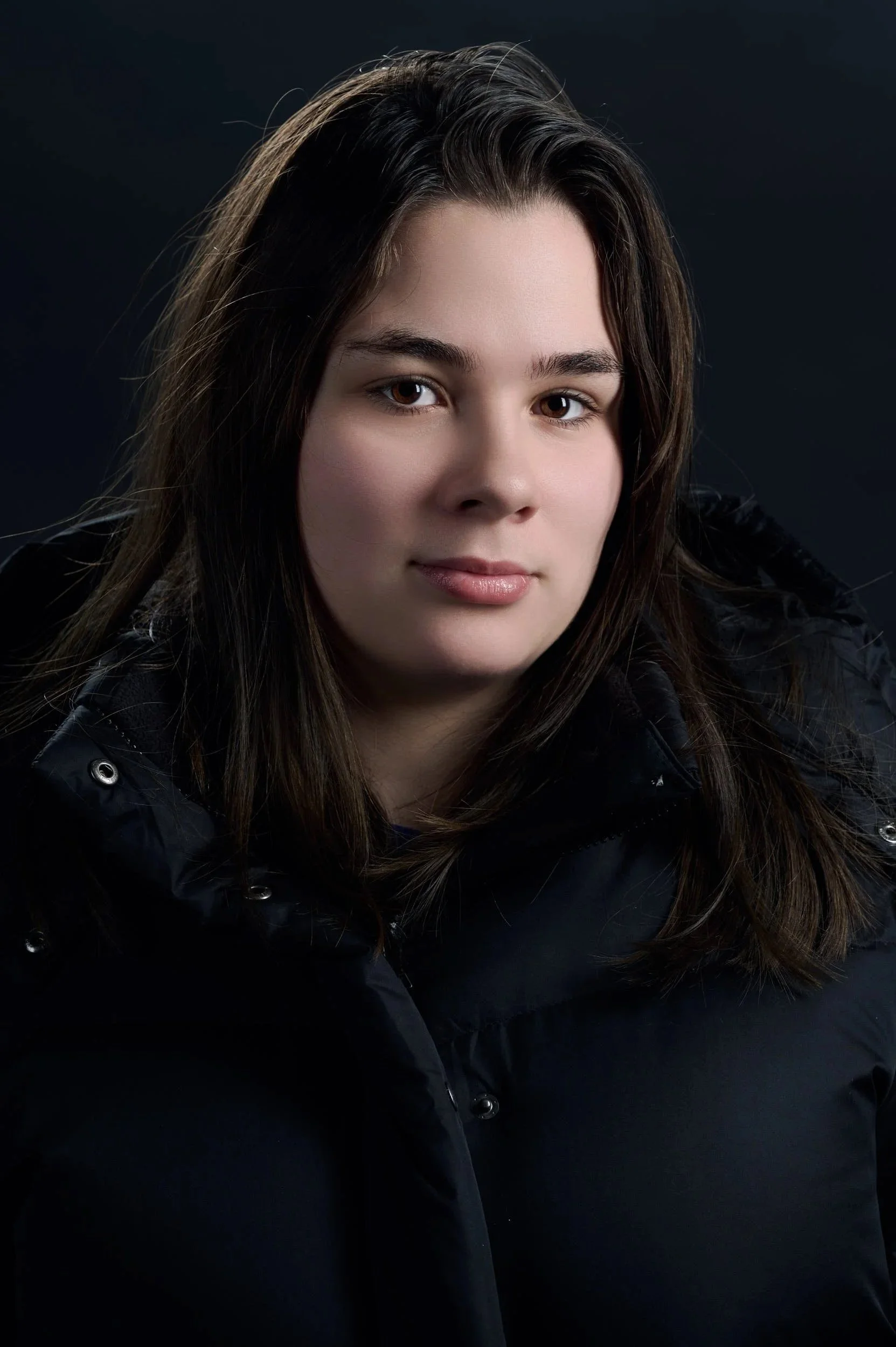 Portrait of a young woman with brown hair wearing a black jacket, against a dark background.