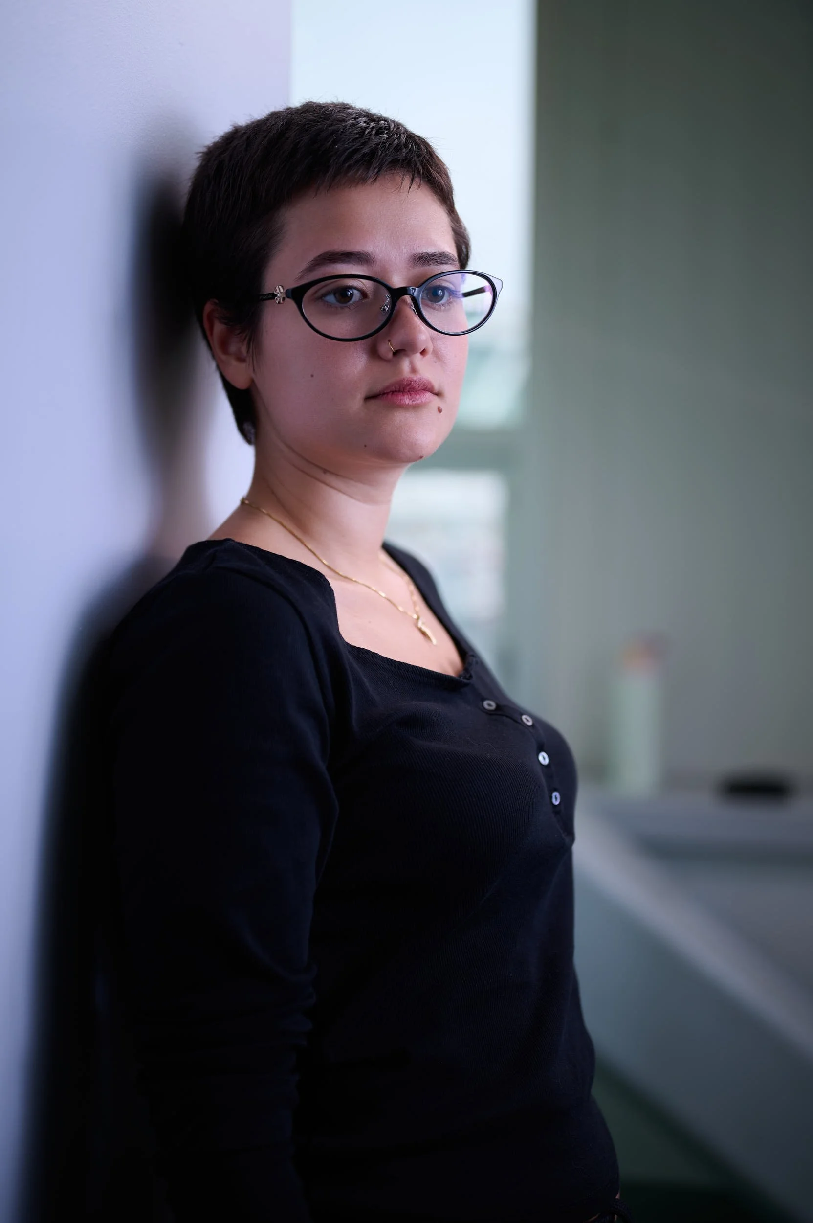 Young woman with short dark hair, glasses, and a nose ring, leaning against a wall inside a building, wearing a black top and a gold necklace.