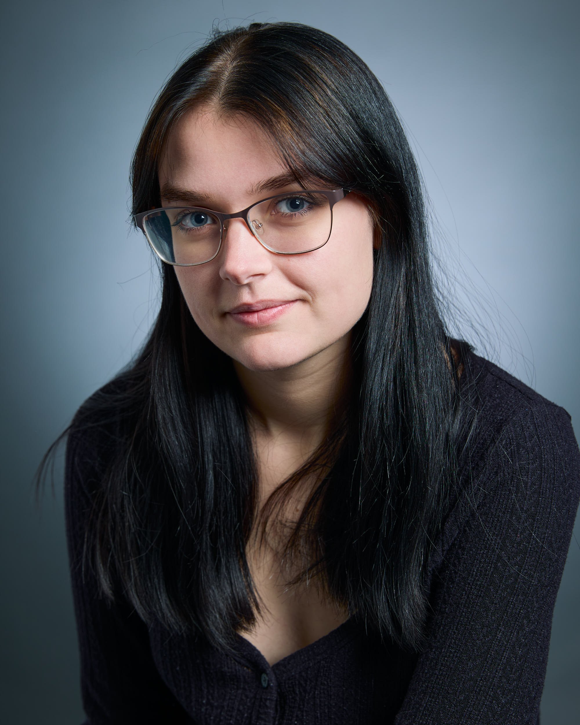 A young woman with long black hair, glasses, and light skin, wearing a dark top, looking at the camera with a slight smile against a gray background.