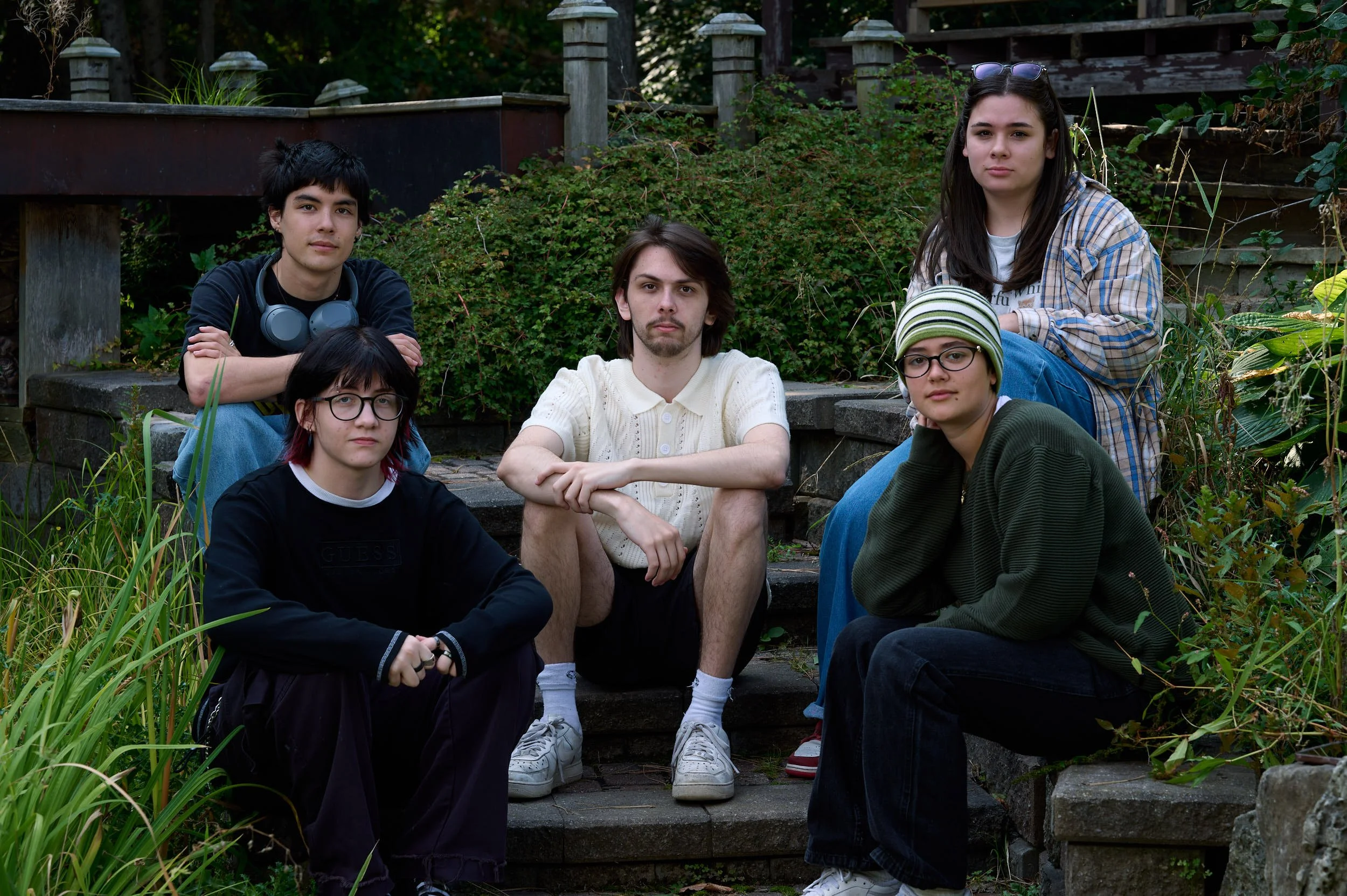Six young people sitting outdoors on stone steps and surrounding greenery, facing the camera with neutral expressions.