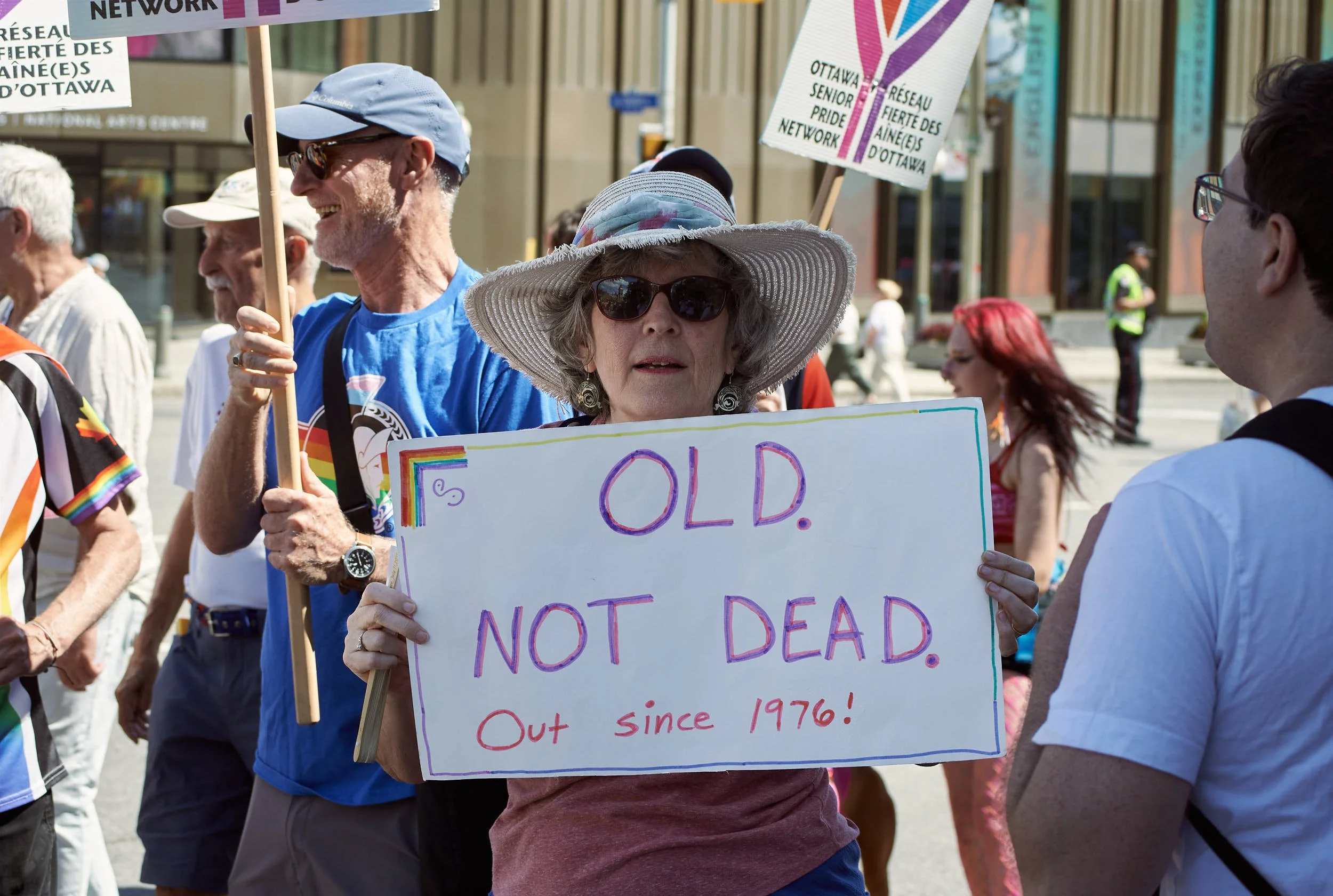 Older woman holding a protest sign that reads 'OLD. NOT DEAD. Out since 1976!' wearing a wide-brimmed hat and sunglasses, participating in a pride parade with people and signs in the background.