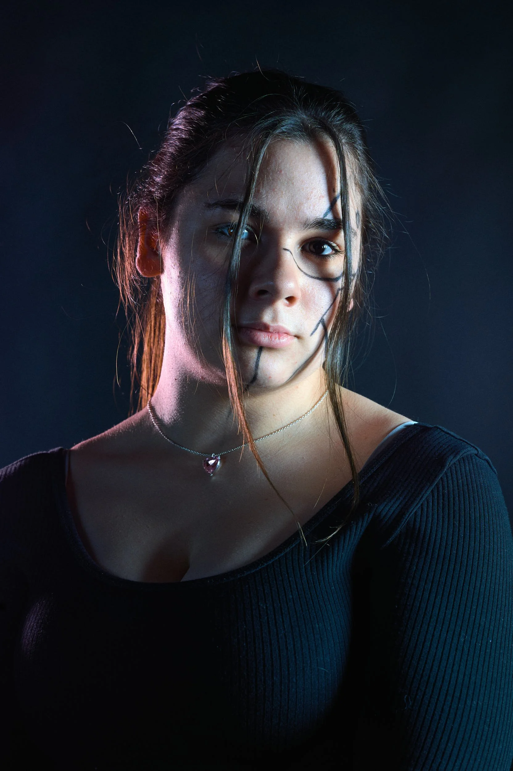 A young woman with multicolored lighting and black hair, wearing a black top and a delicate necklace, looking directly at the camera.