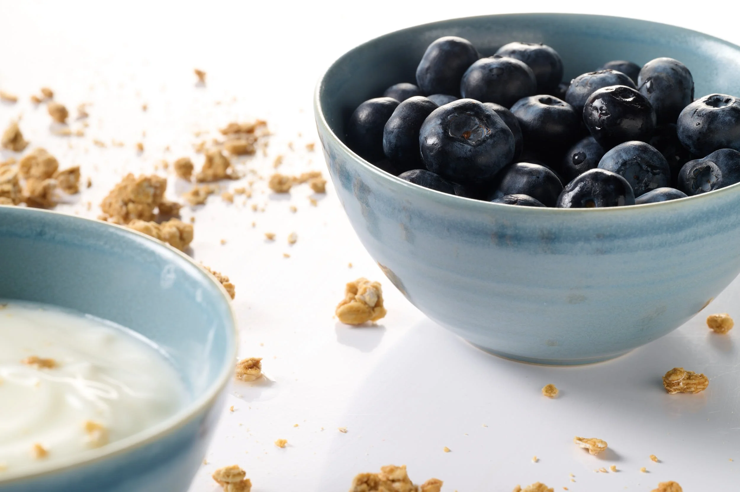 Blue ceramic bowls, one filled with blueberries and the other with yogurt, on a white surface with scattered granola crumbs.