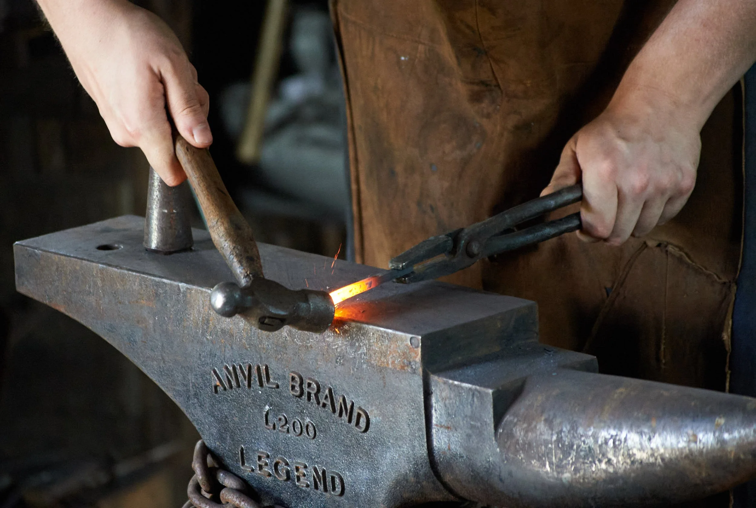 A blacksmith heating a metal piece in a forge, holding tongs with one hand and a hammer with the other.