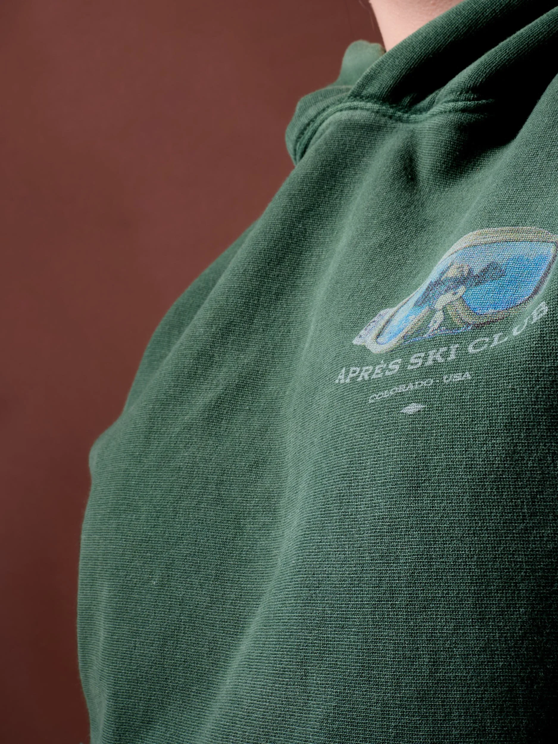 Close-up of a green Patagonia sweatshirt with an embroidered logo and text reading "Après Ski Club, Colorado, USA" on the chest, against a brown background.