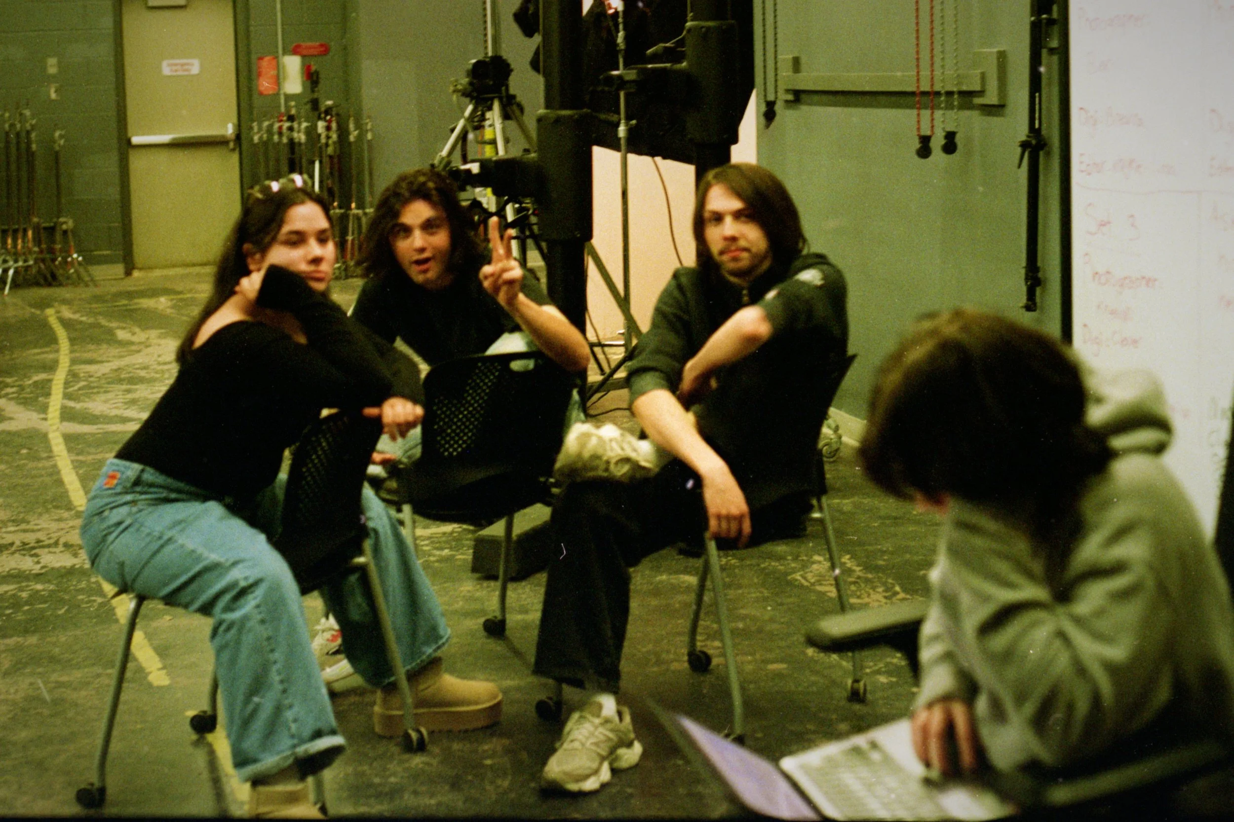 Four young people sitting on chairs in a room with equipment, one person with long hair making a peace sign, another reading a laptop, and others engaged in a casual conversation or activity.