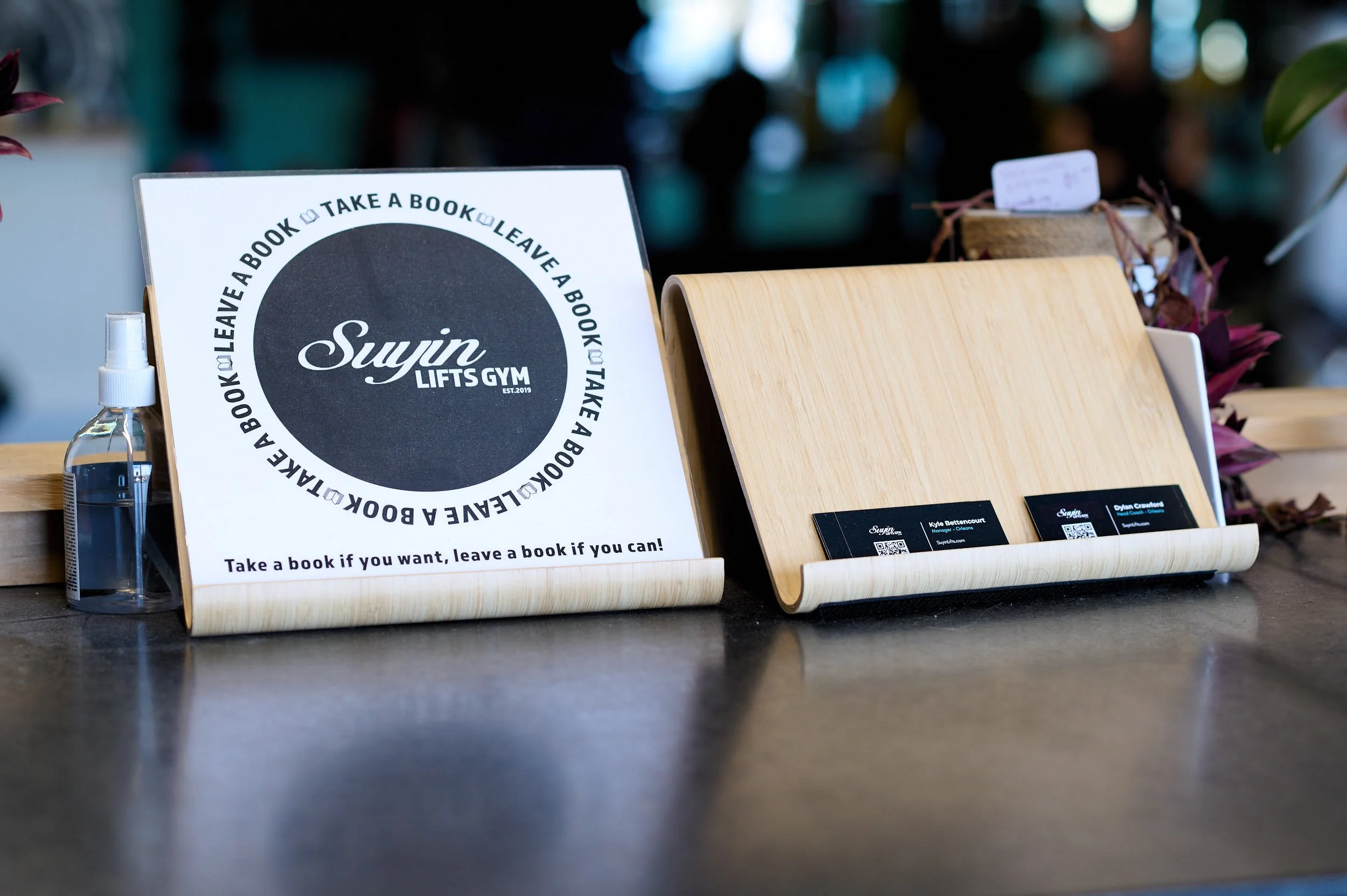 A wooden table with a sign promoting a book exchange program at Suyn Lift Gym, with a spray bottle nearby and a wooden stand holding business cards.