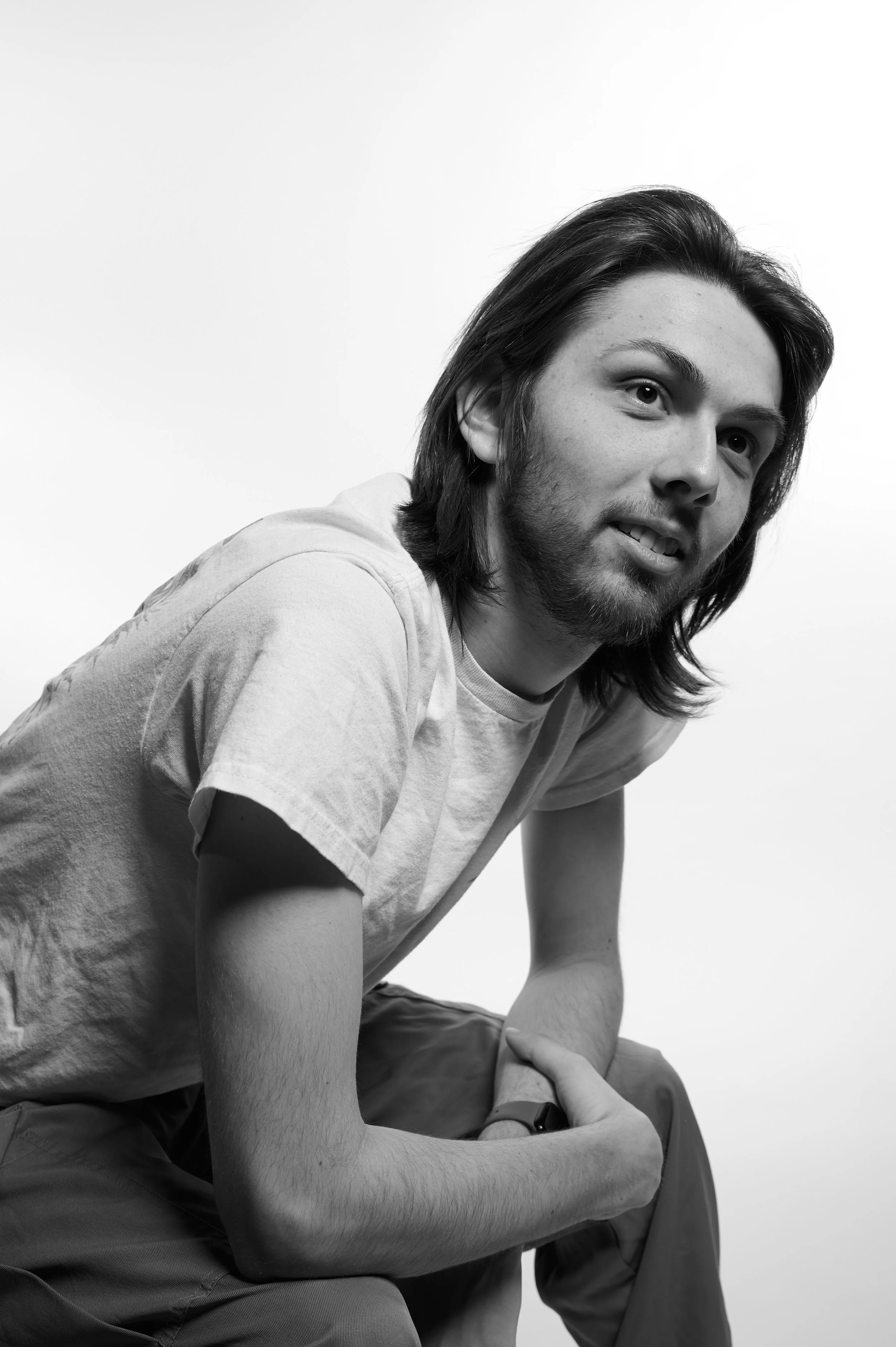 Black-and-white photo of a young man with long hair and a beard, wearing a t-shirt and pants, sitting with his elbow on his knee and looking slightly to the right.