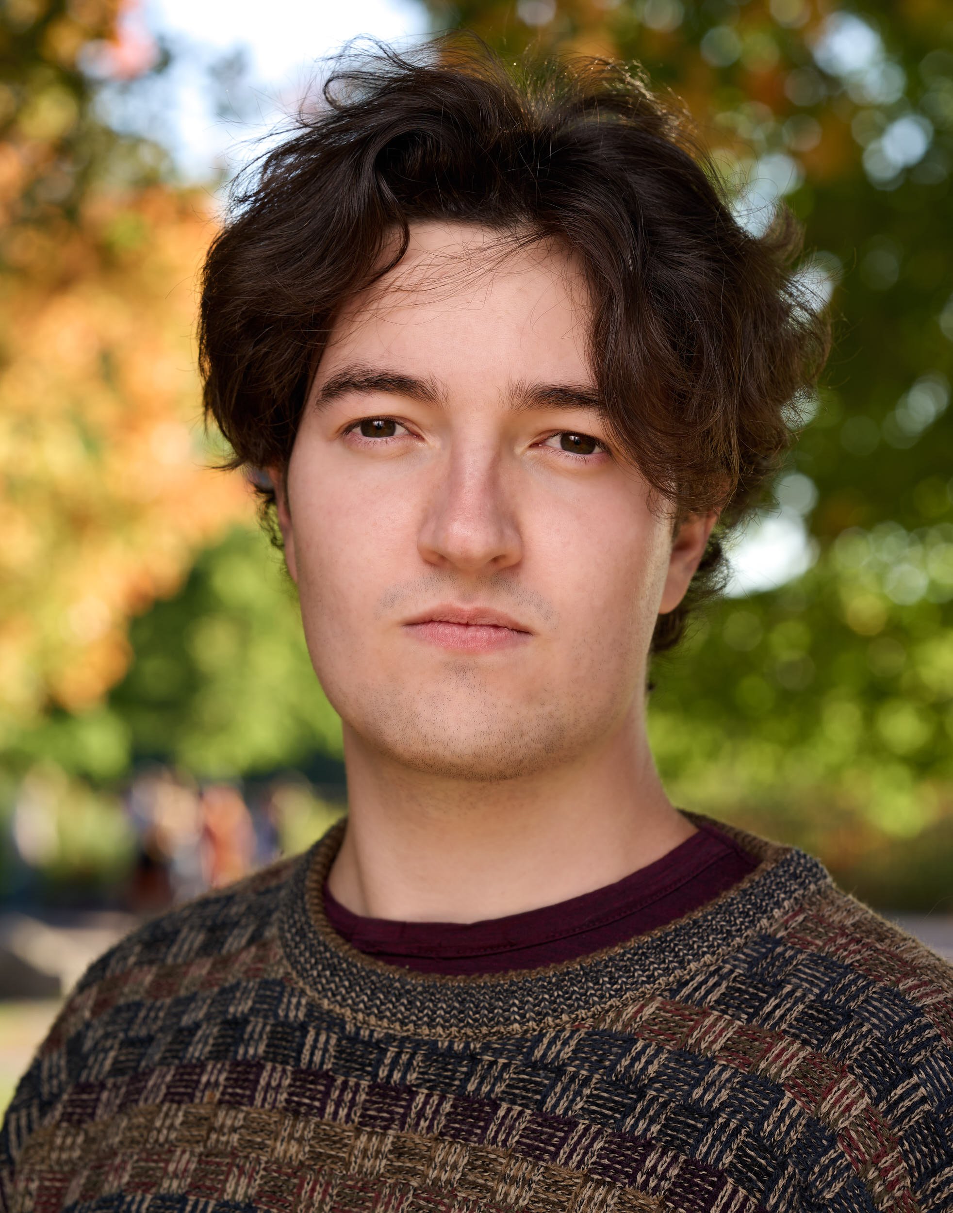 A young man with dark curly hair and light skin looking slightly off camera in an outdoor setting with autumn trees in the background.