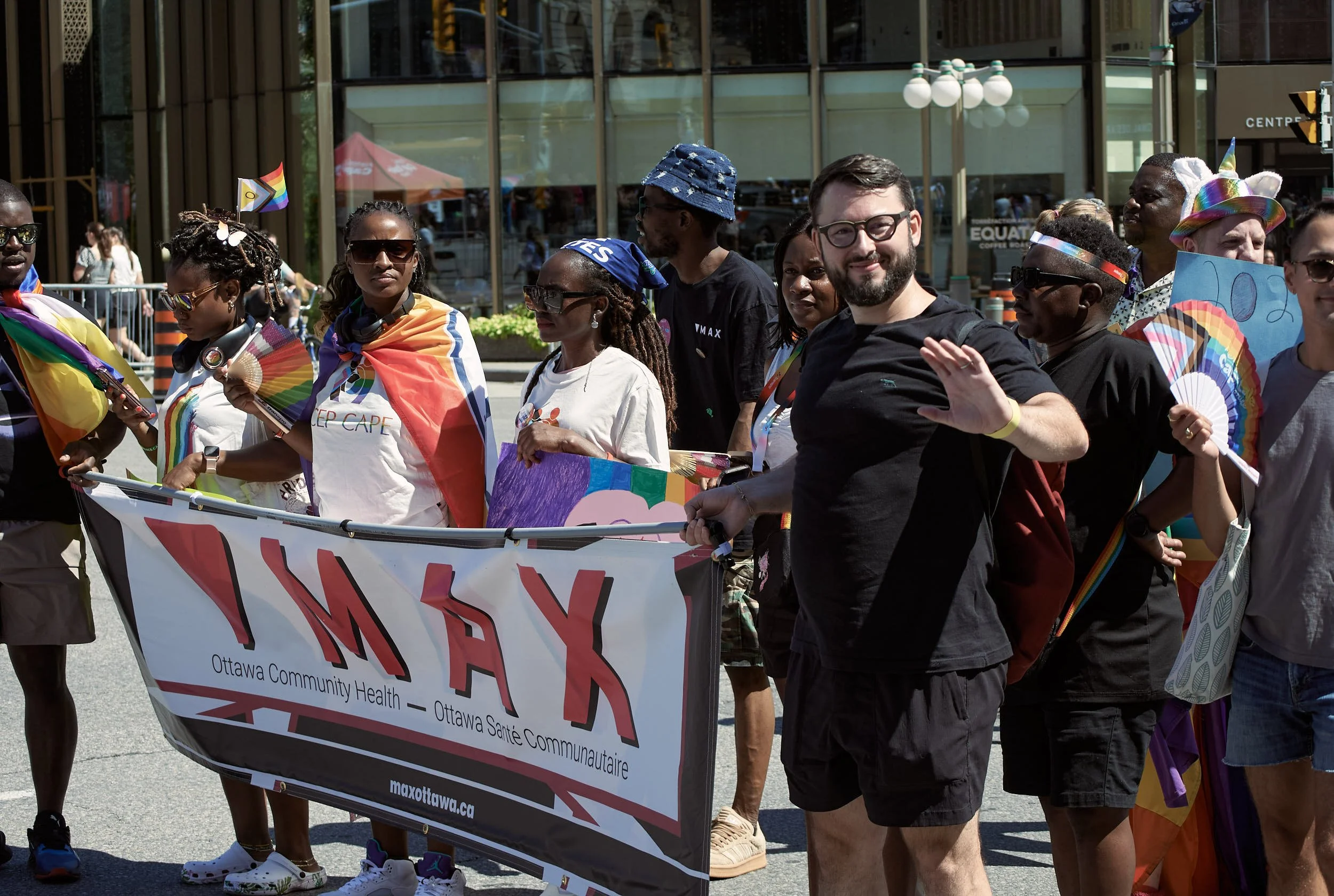 A diverse group of people participating in a pride parade, holding a banner that reads "Ottawa Community Health," with some wearing rainbow-themed accessories and flags, walking in the street on a sunny day.