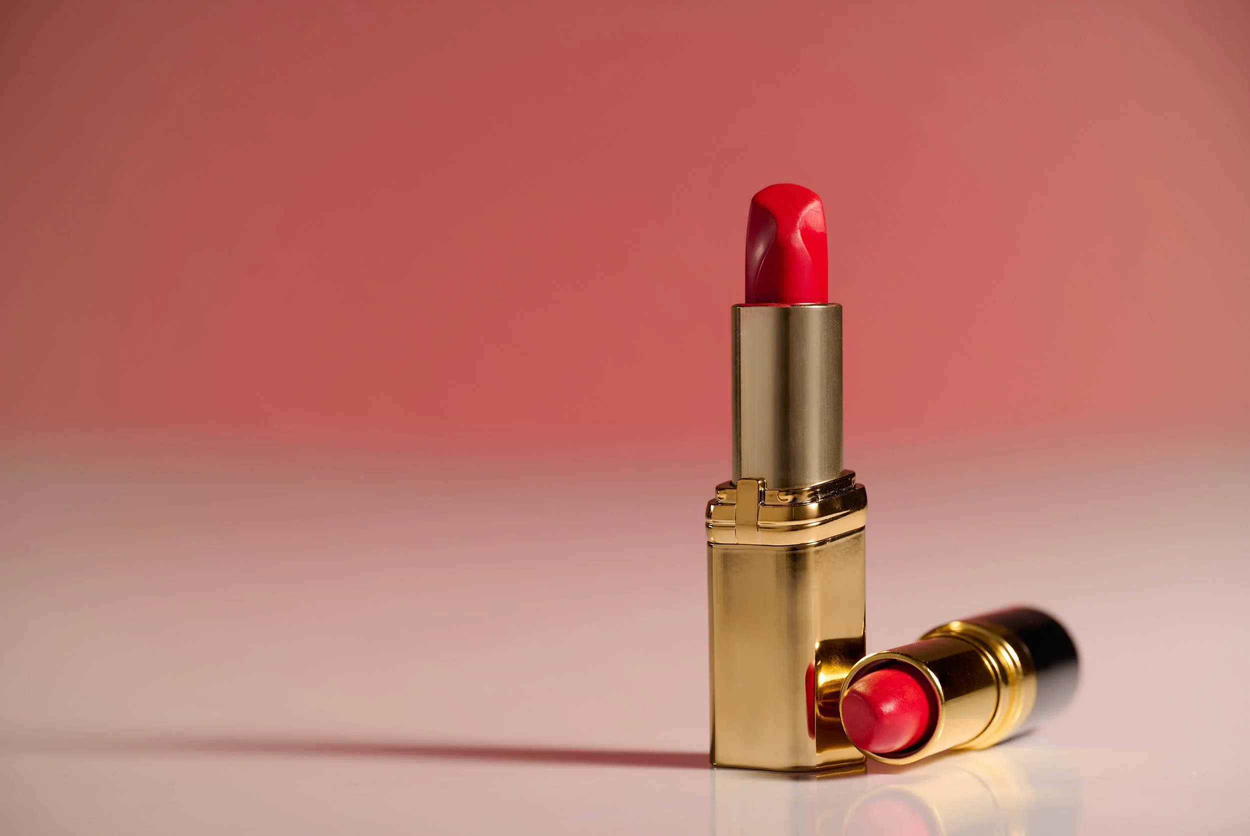 A gold lipstick case with a red lipstick inside and another lipstick lying on its side in front of it, both against a pink gradient background.