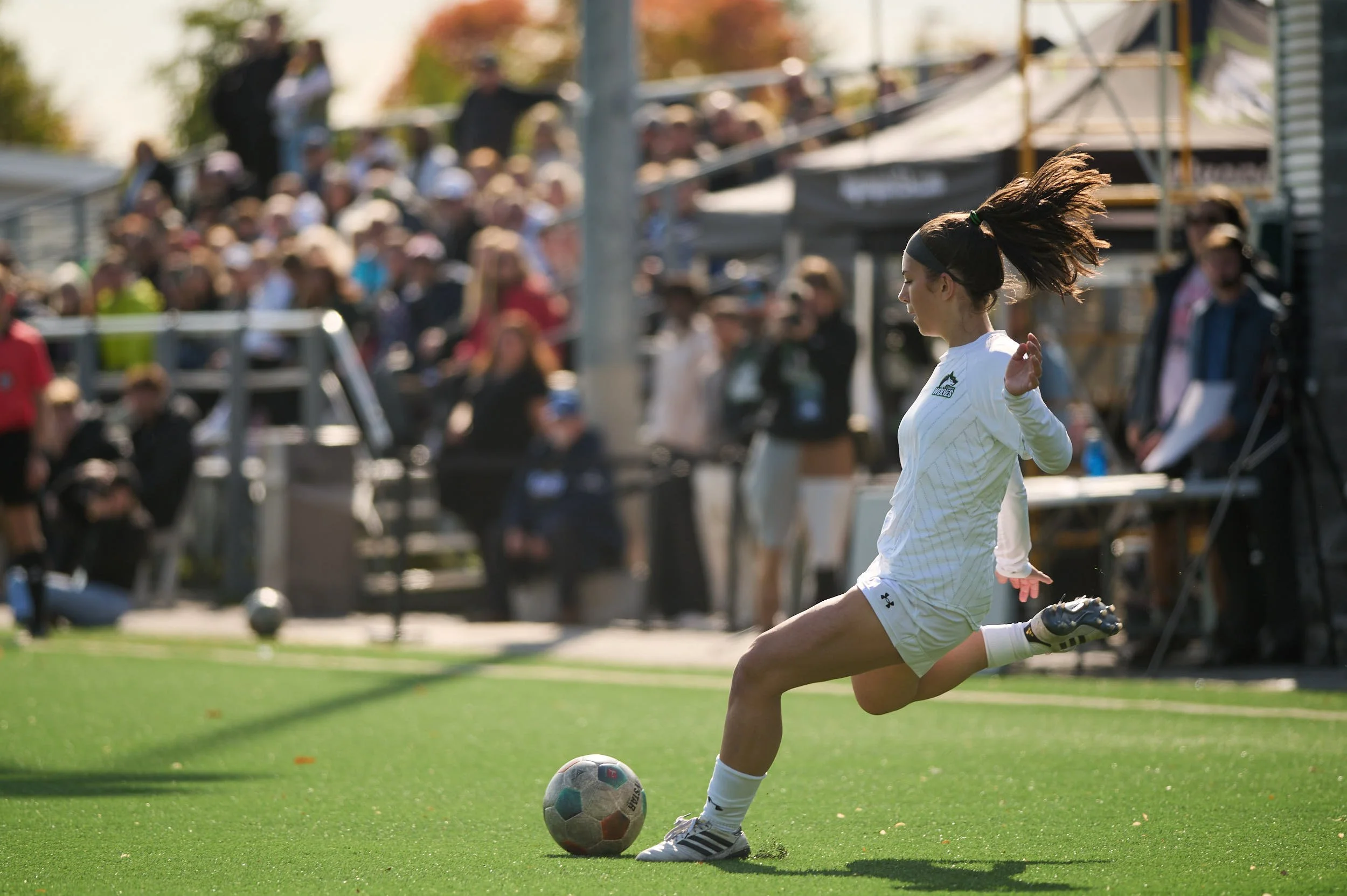 A female soccer player in white uniform kicking a soccer ball on the field during a game.