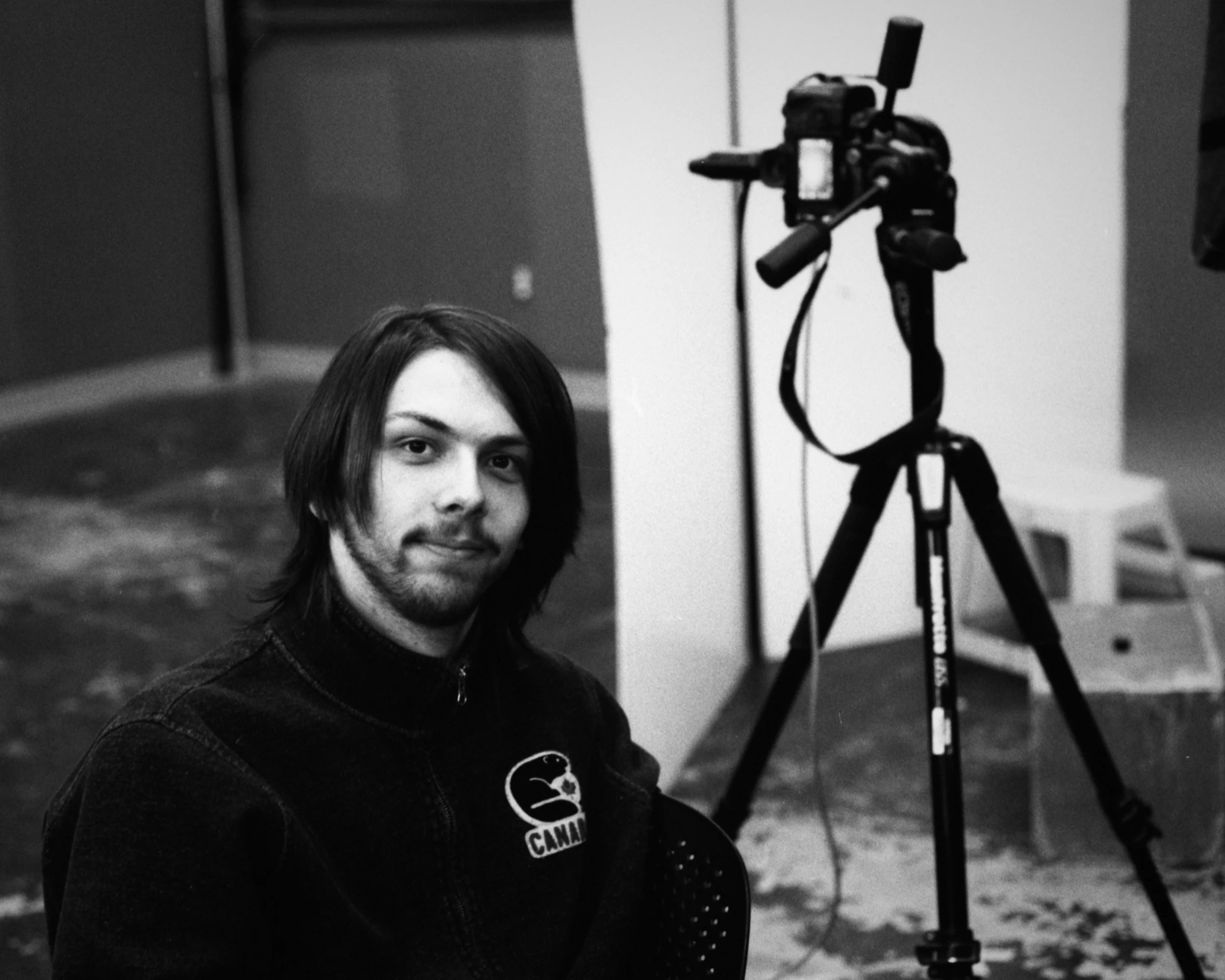 A young man with medium-length hair and a goatee sitting in front of a camera on a tripod in a room.