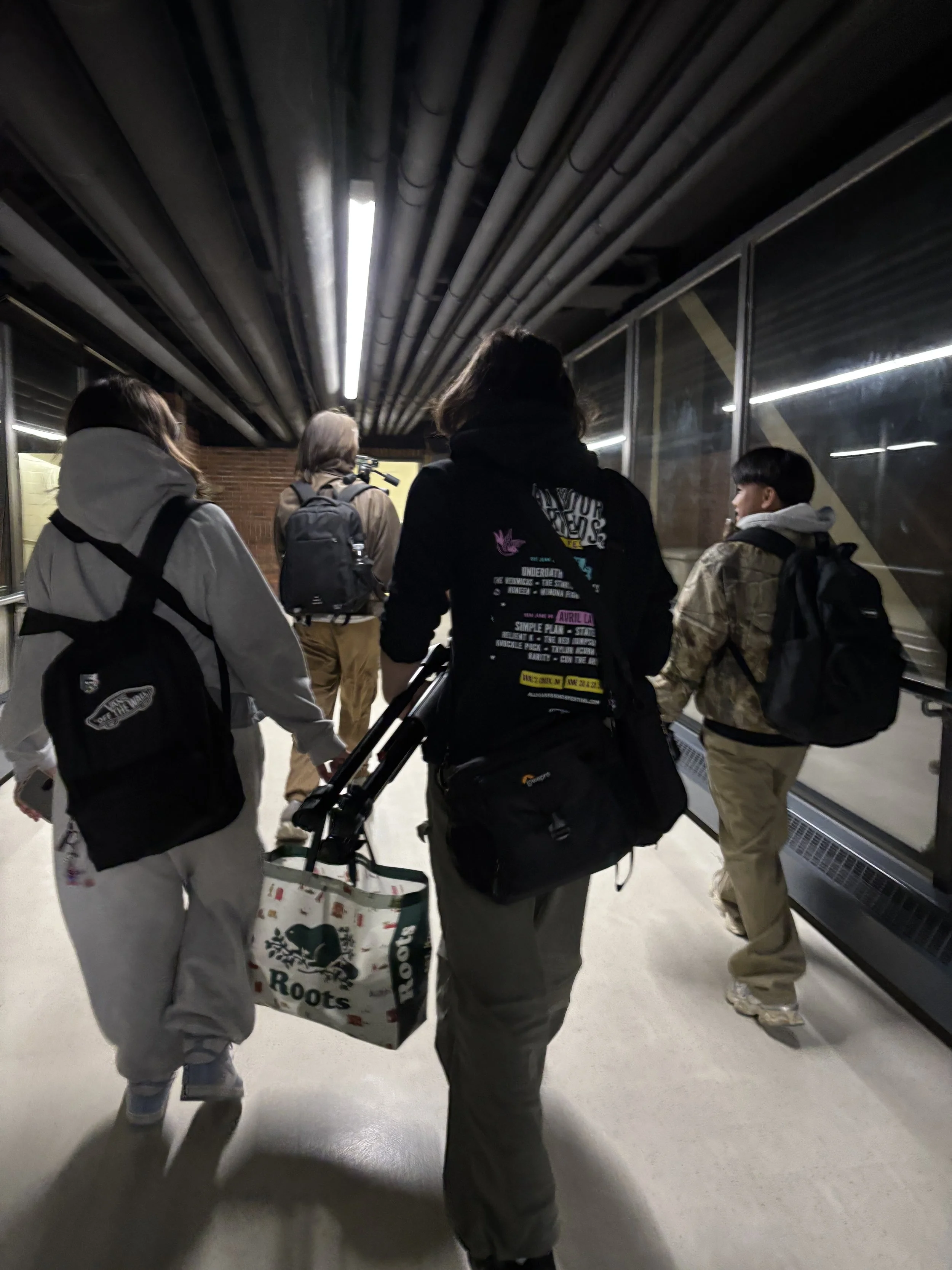 A group of five people walking in an underground passageway, carrying backpacks and shopping bags, with a glass window on the right and ceiling pipes overhead.